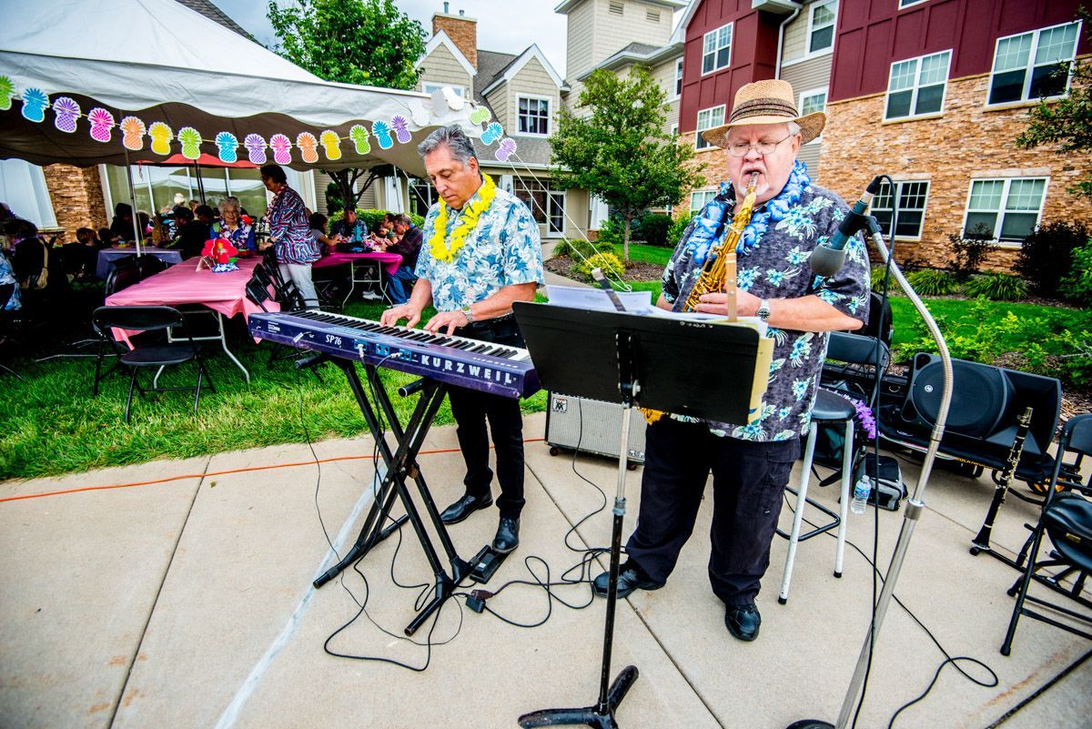 Two musicians play a keyboard and saxophone on an outdoor patio in front of a party under a white tent.