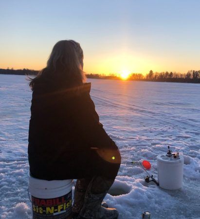 A person sits on a bucket ice fishing on a frozen lake at sunset.