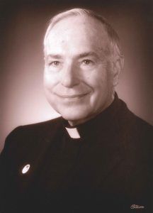 A portrait of a clergyman in a dark suit with a clerical collar, posing with a gentle smile against a soft brown background.