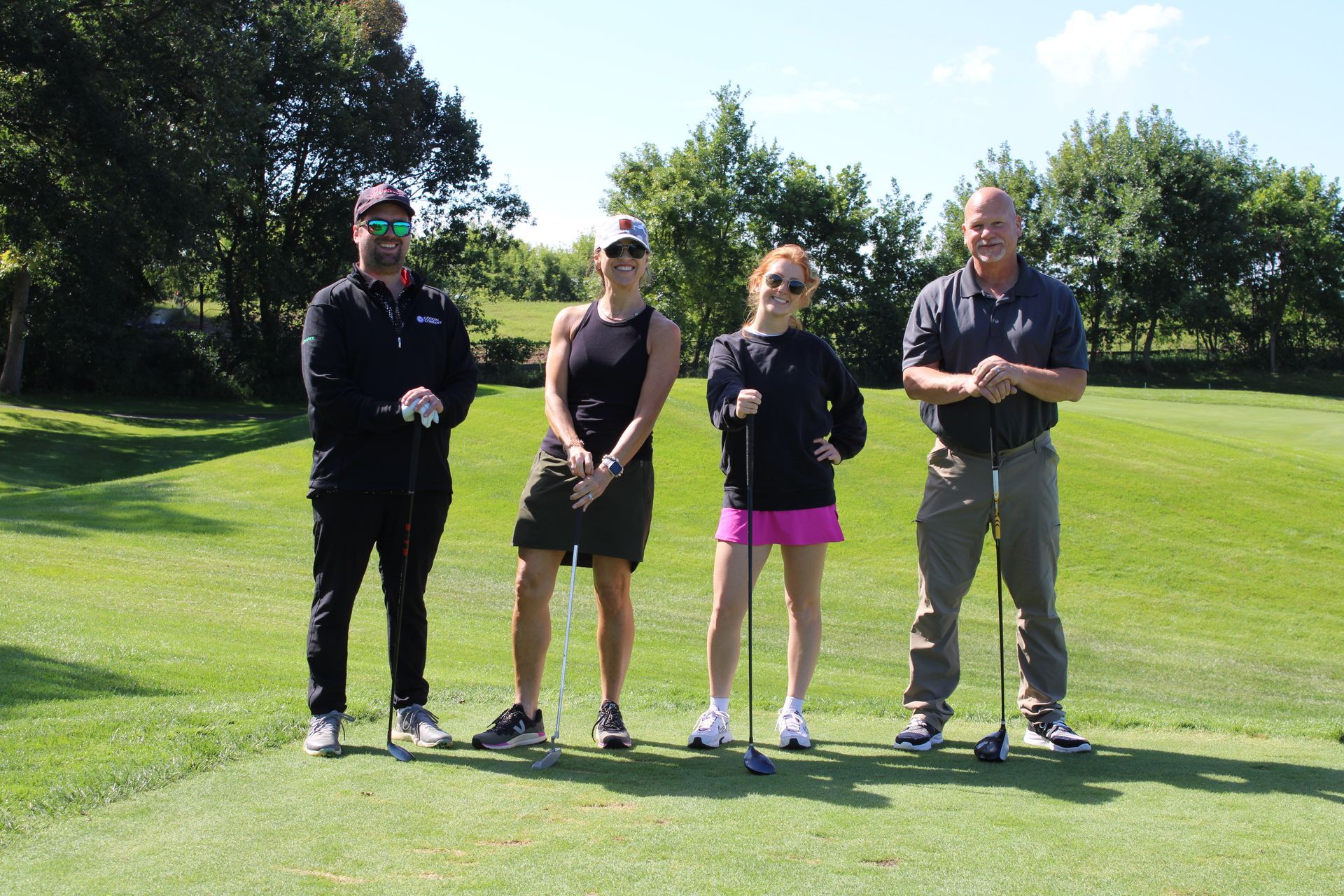 Four people stand on a grassy golf course, each holding a golf club, posed together outdoors under a sunny sky.