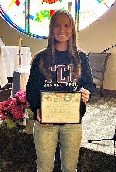 A person in a TCU sweatshirt smiles while holding an award certificate indoors in front of a stained-glass window.