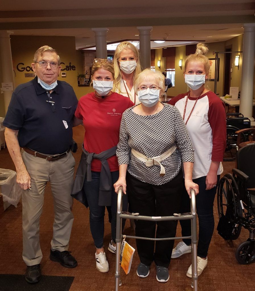 Five people stand together indoors, four wearing face masks, one with a walker in a room with cafe seating.