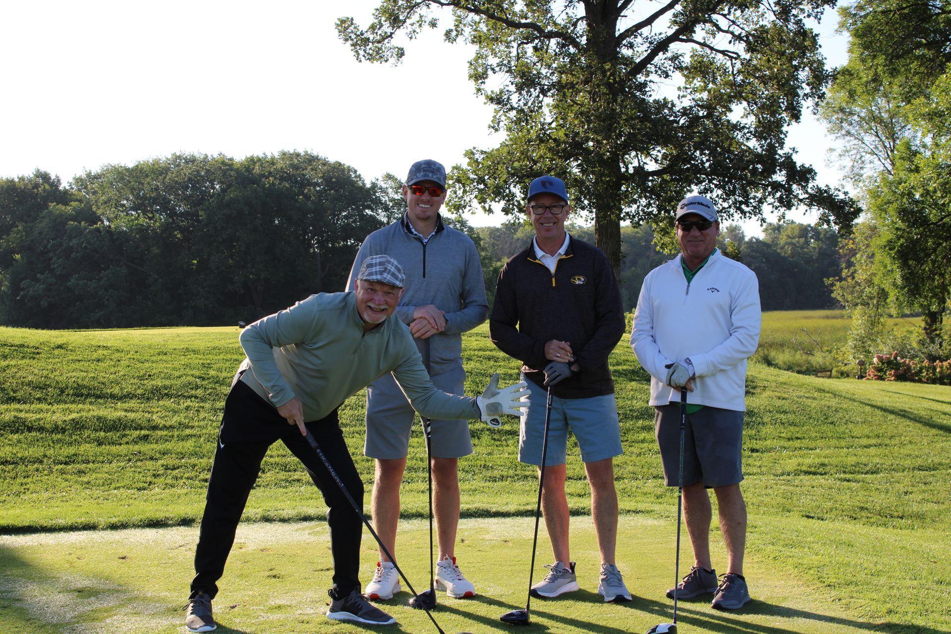 Four golfers in hats and casual golf attire pose together on a sunny, grassy tee box holding their clubs.
