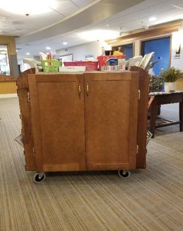 A wooden rolling supply cart with two doors stands in a hallway filled with medical equipment and baskets.