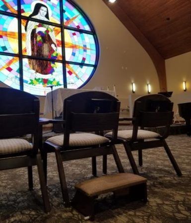Dark chairs and a kneeling bench face a circular stained glass window in a carpeted church sanctuary.