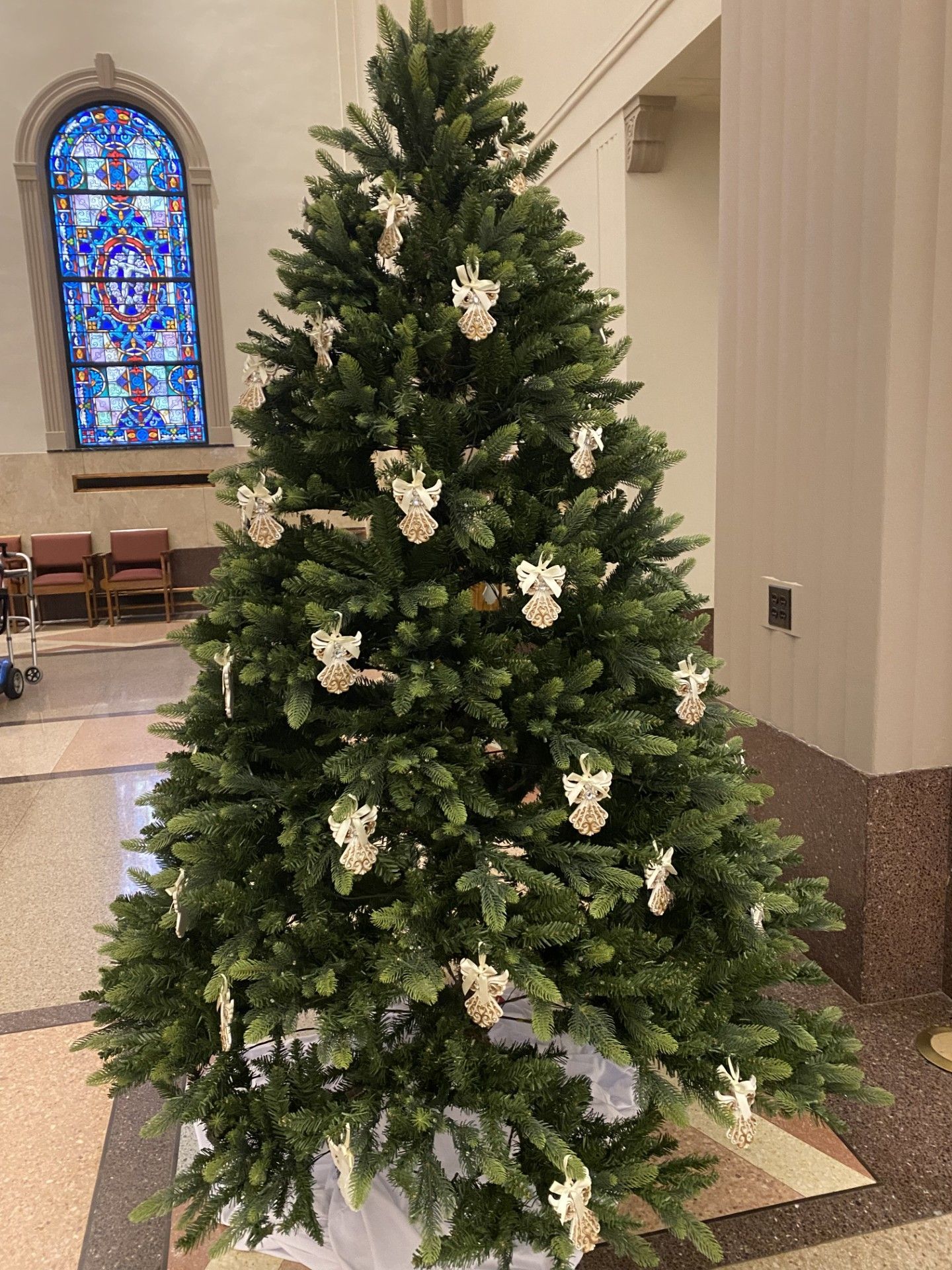 A Christmas tree decorated with white angel ornaments, standing in a hall with a stained glass window in the background.