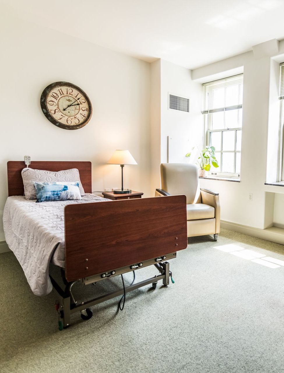 A room with a single hospital bed, bedside lamp, a tan armchair, and a wall clock, featuring light walls and carpeting.