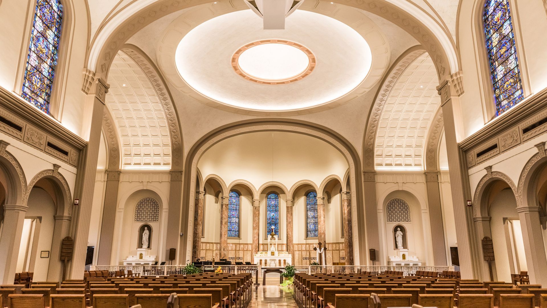 The interior of a grand church with rows of wooden pews facing an altar featuring stained-glass windows and arches.