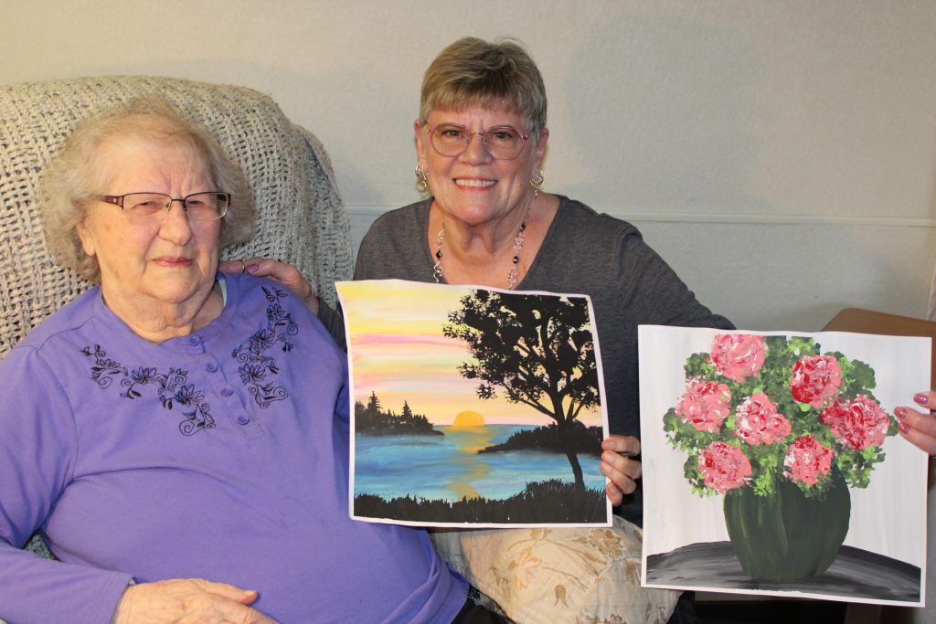 Two people sit side-by-side, holding up their completed paintings of a sunset landscape and a bouquet of flowers.