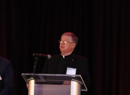 A person in clerical clothing stands at a podium and speaks into a microphone against a dark curtain background.