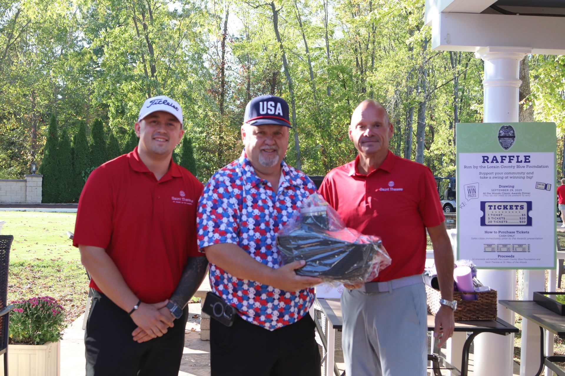 Three individuals stand outdoors, with the center person holding a gift, next to a raffle sign at a golf tournament.