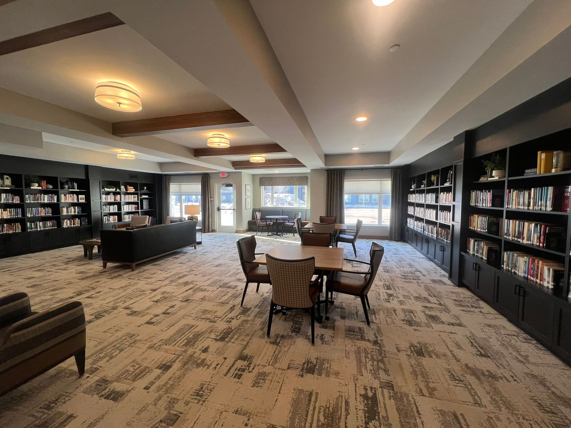 A spacious library with bookshelves lining the walls, a central dining table with chairs, and soft carpeted flooring.