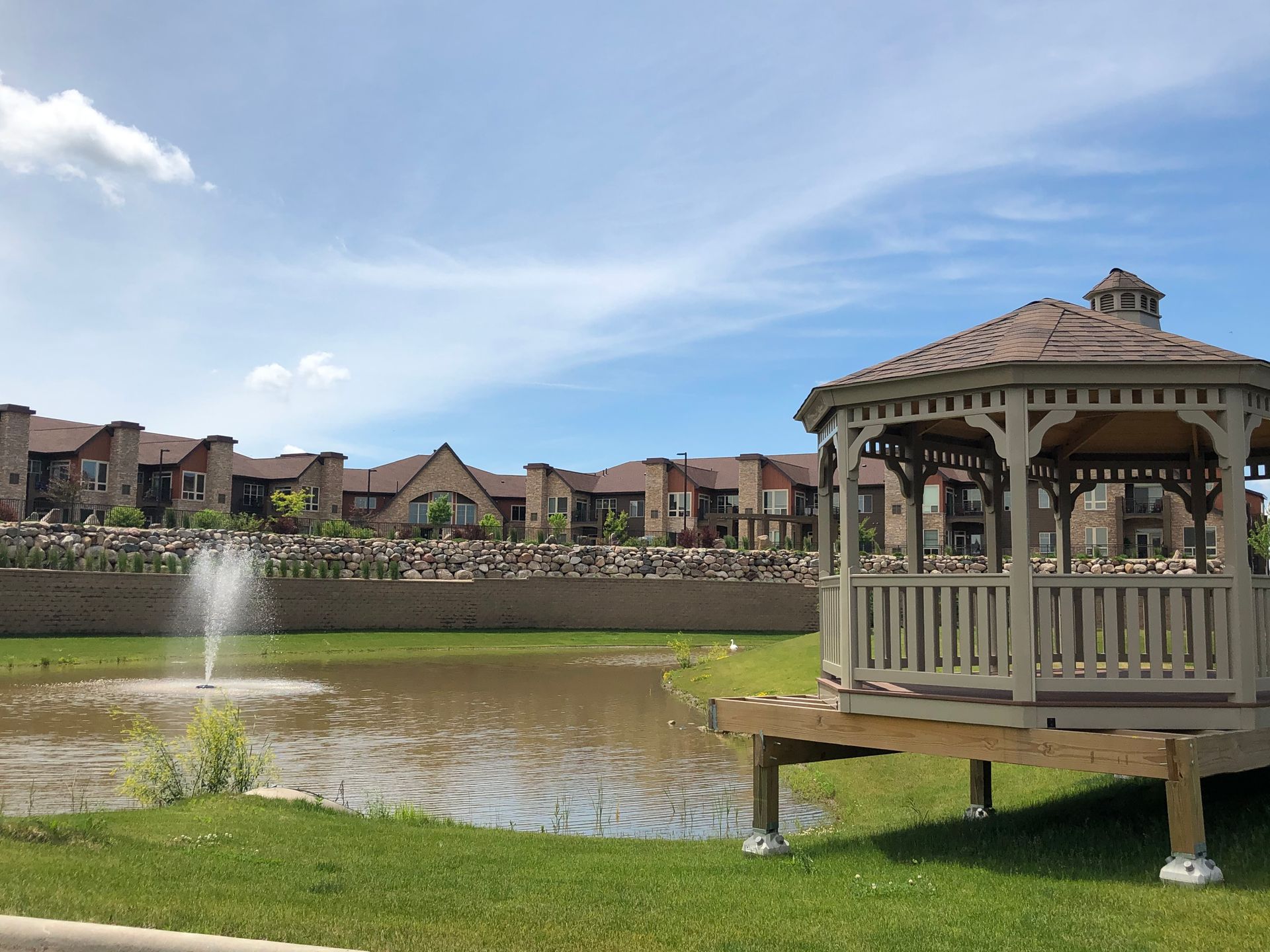 A tan wooden gazebo overlooks a small pond with a water fountain, set against a background of apartment buildings.