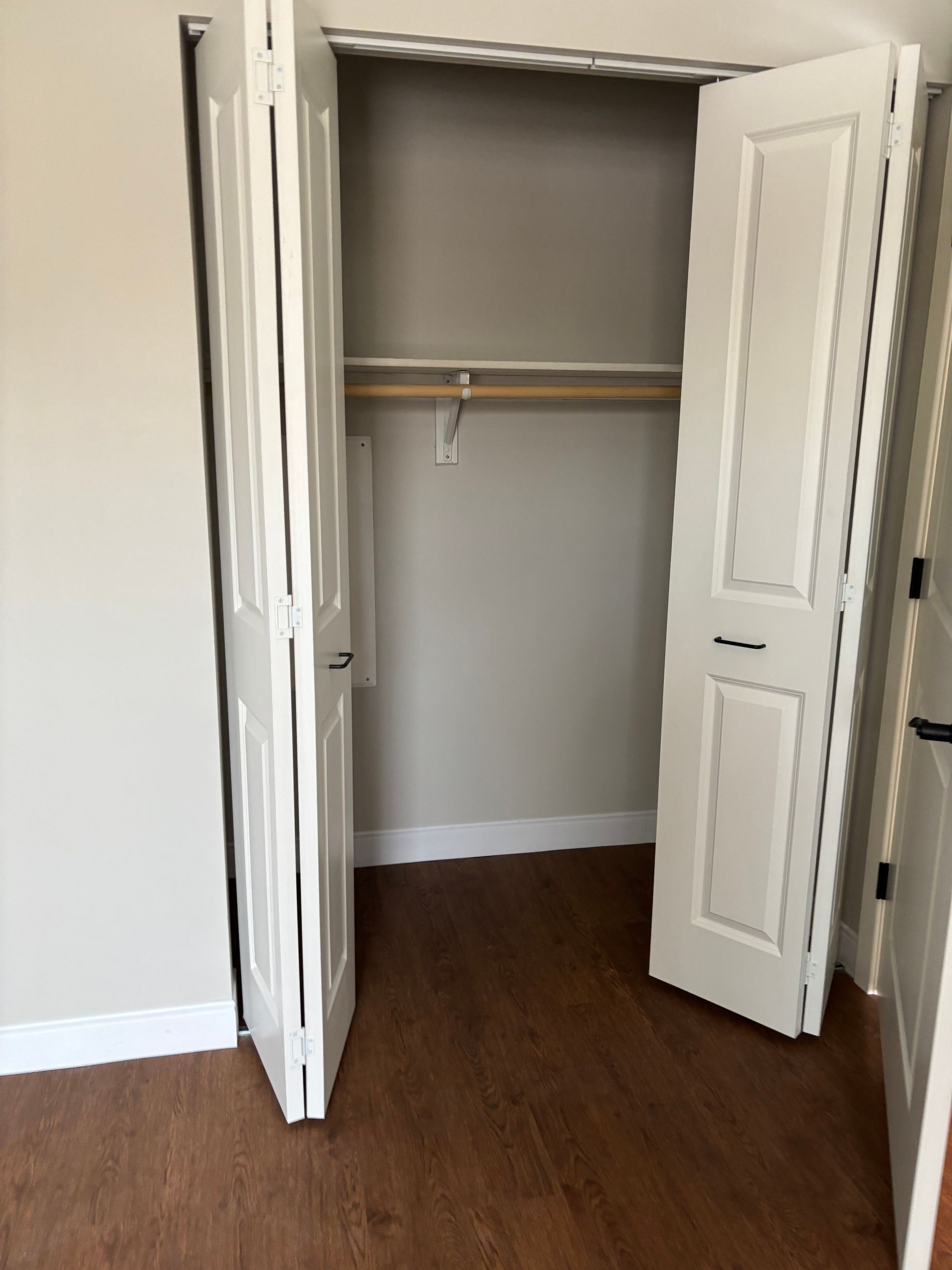 An open beige closet with white bi-fold doors, a wooden hanging rod, and wood-look flooring.
