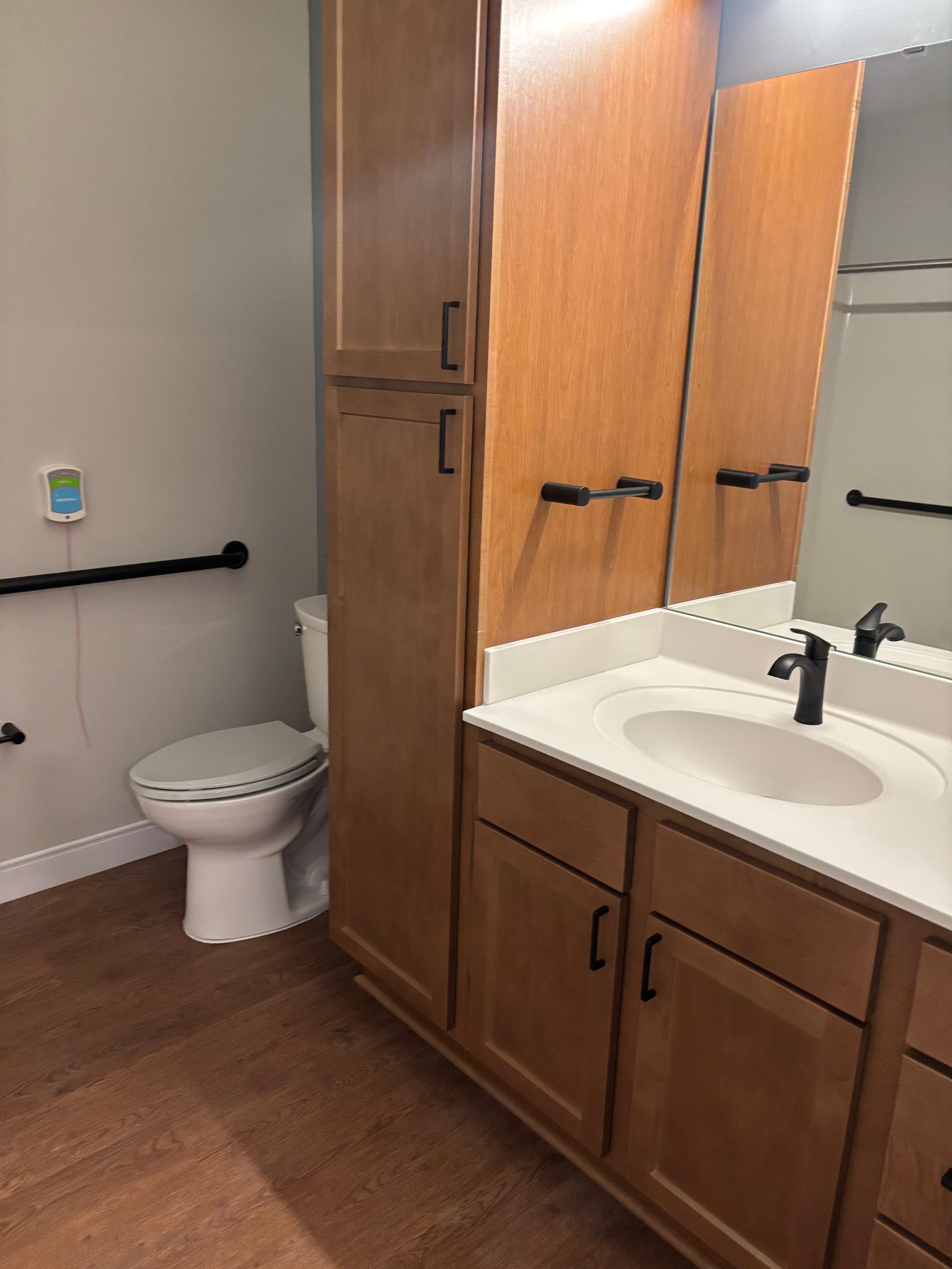 Accessible bathroom with a wooden vanity, white sink, and a toilet equipped with a black wall-mounted support grab bar.
