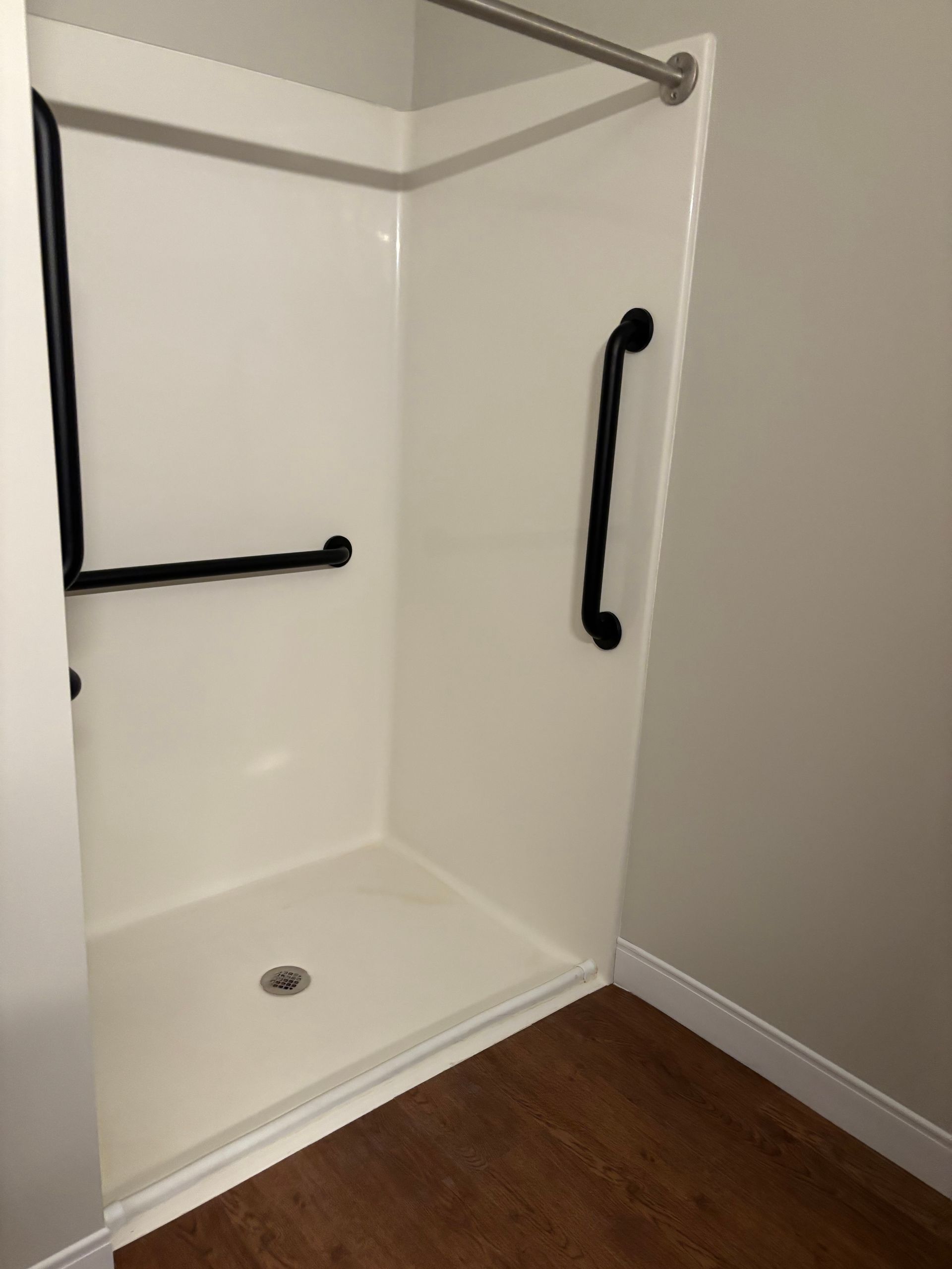 A white walk-in shower stall with black grab bars installed on the side and back walls, set against a light-colored wall.
