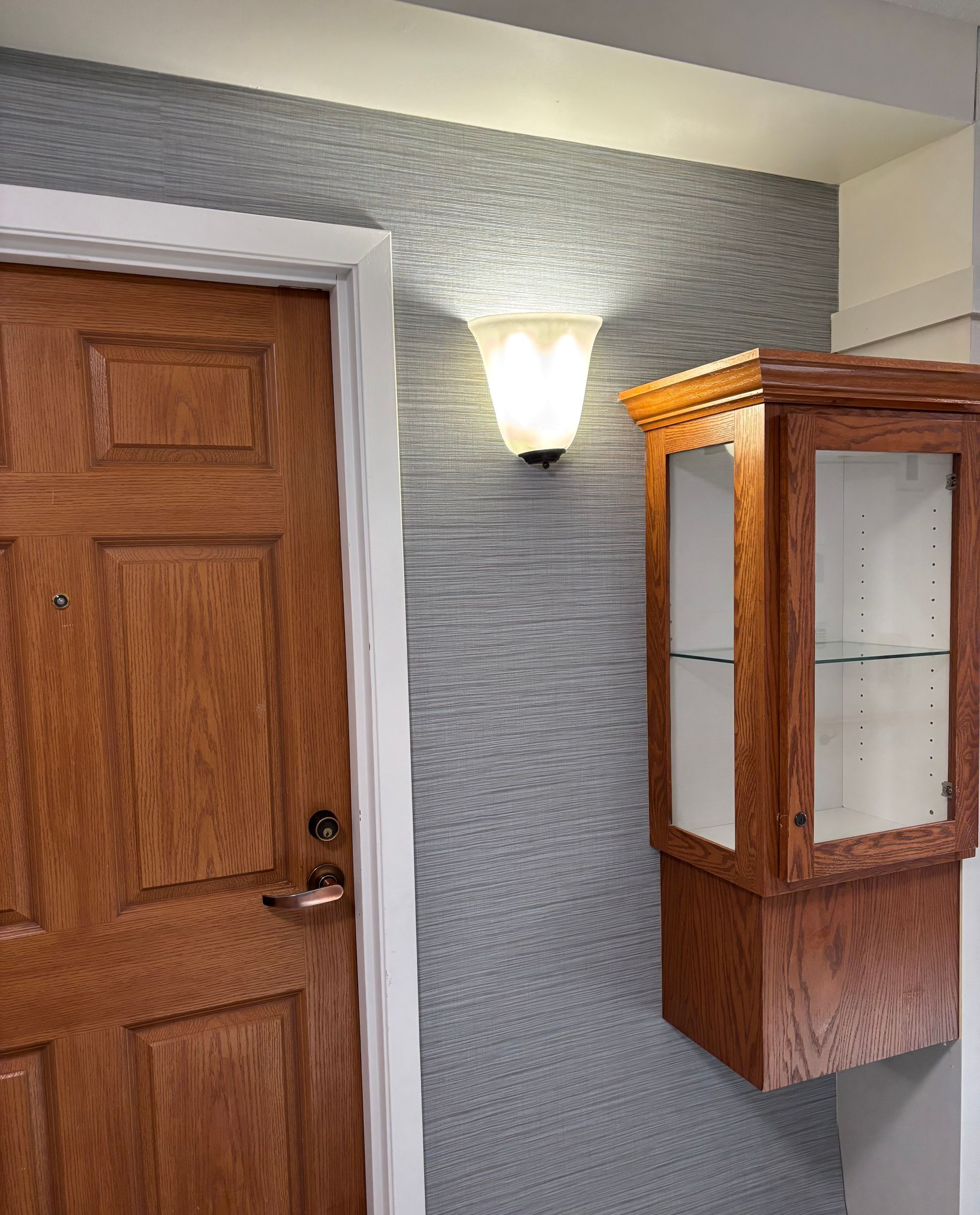 A wooden door with a brass handle next to a wall with textured grey wallpaper, a wall sconce, and a wood-framed cabinet.