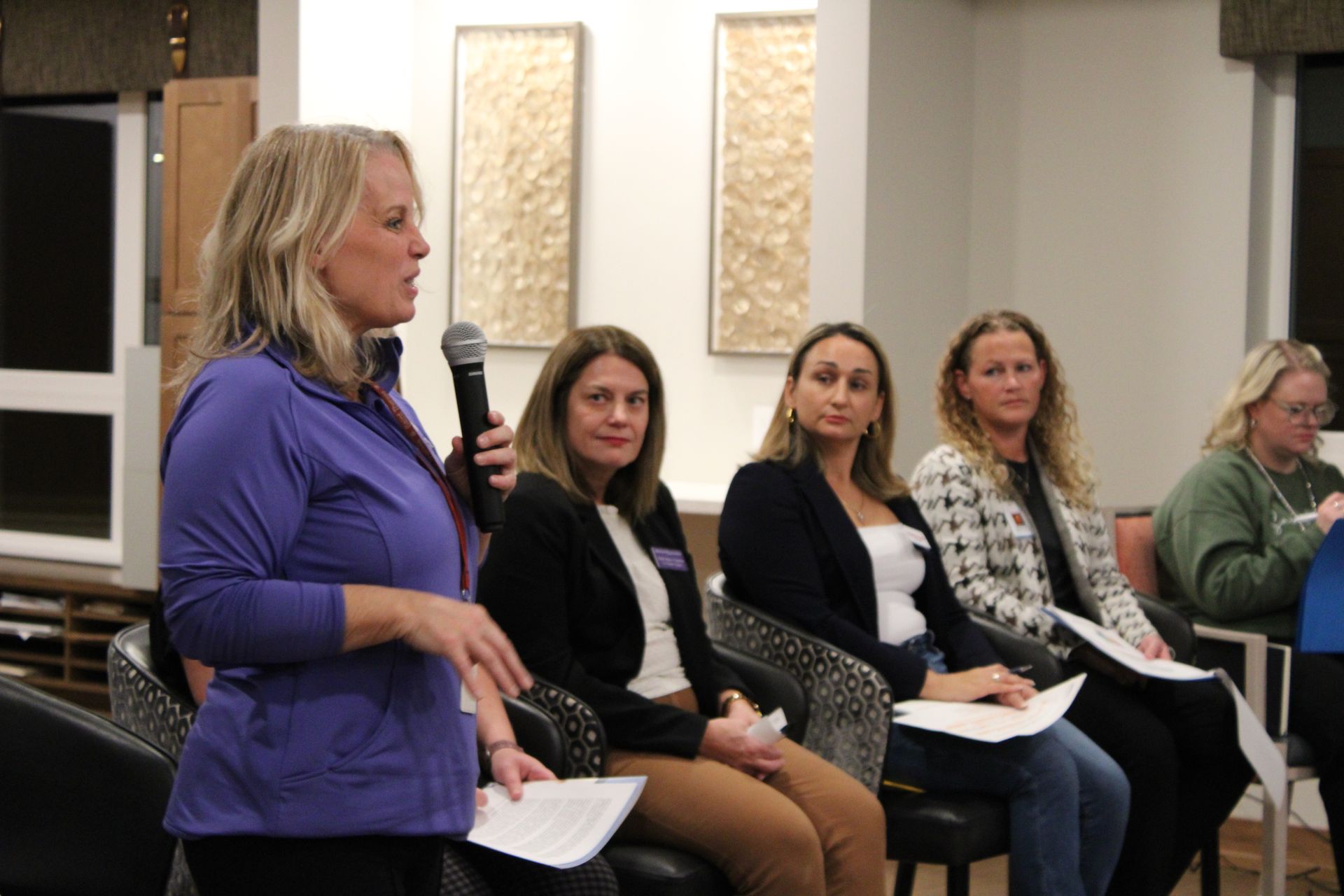 A woman in a purple jacket speaks into a microphone before a panel of four seated people in an indoor room.