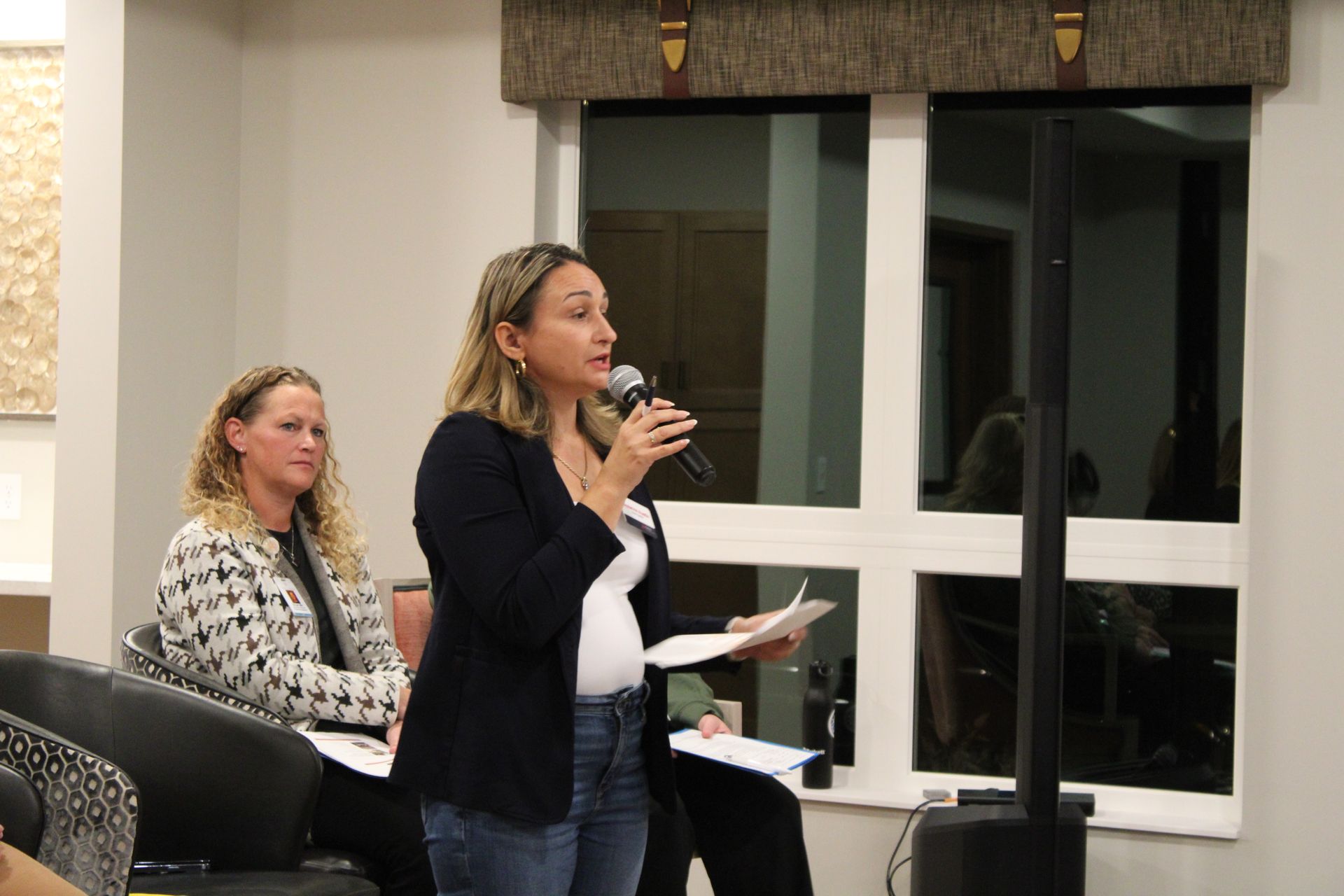 A person speaks into a microphone while holding a paper, as another person sits and listens in a lit indoor room.