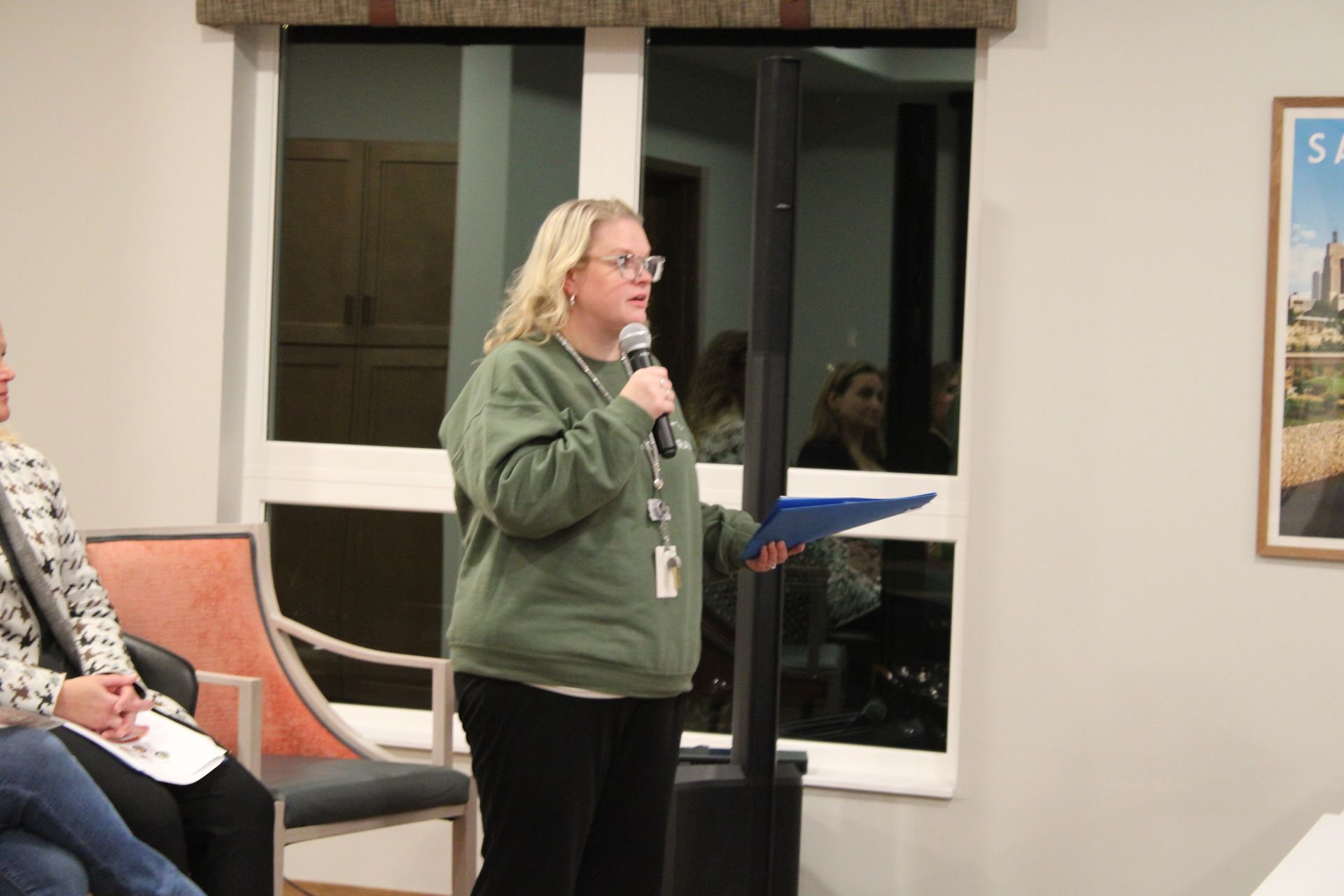 A speaker in a green sweatshirt holding a microphone and blue folder stands in front of a window at an indoor event.
