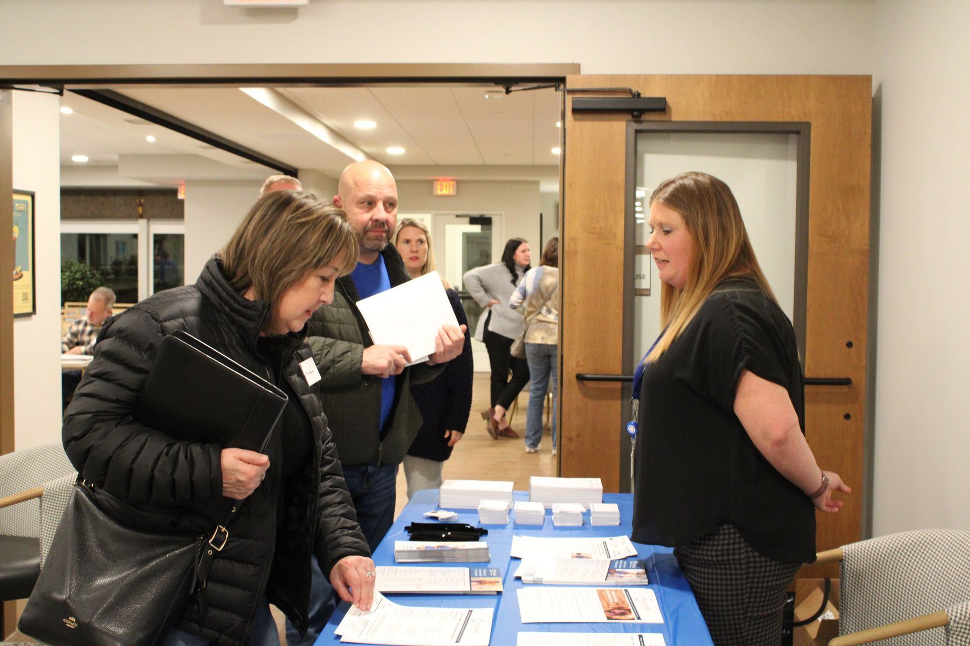 Three adults interact at an information table in a bright hallway, with others visible in the background.