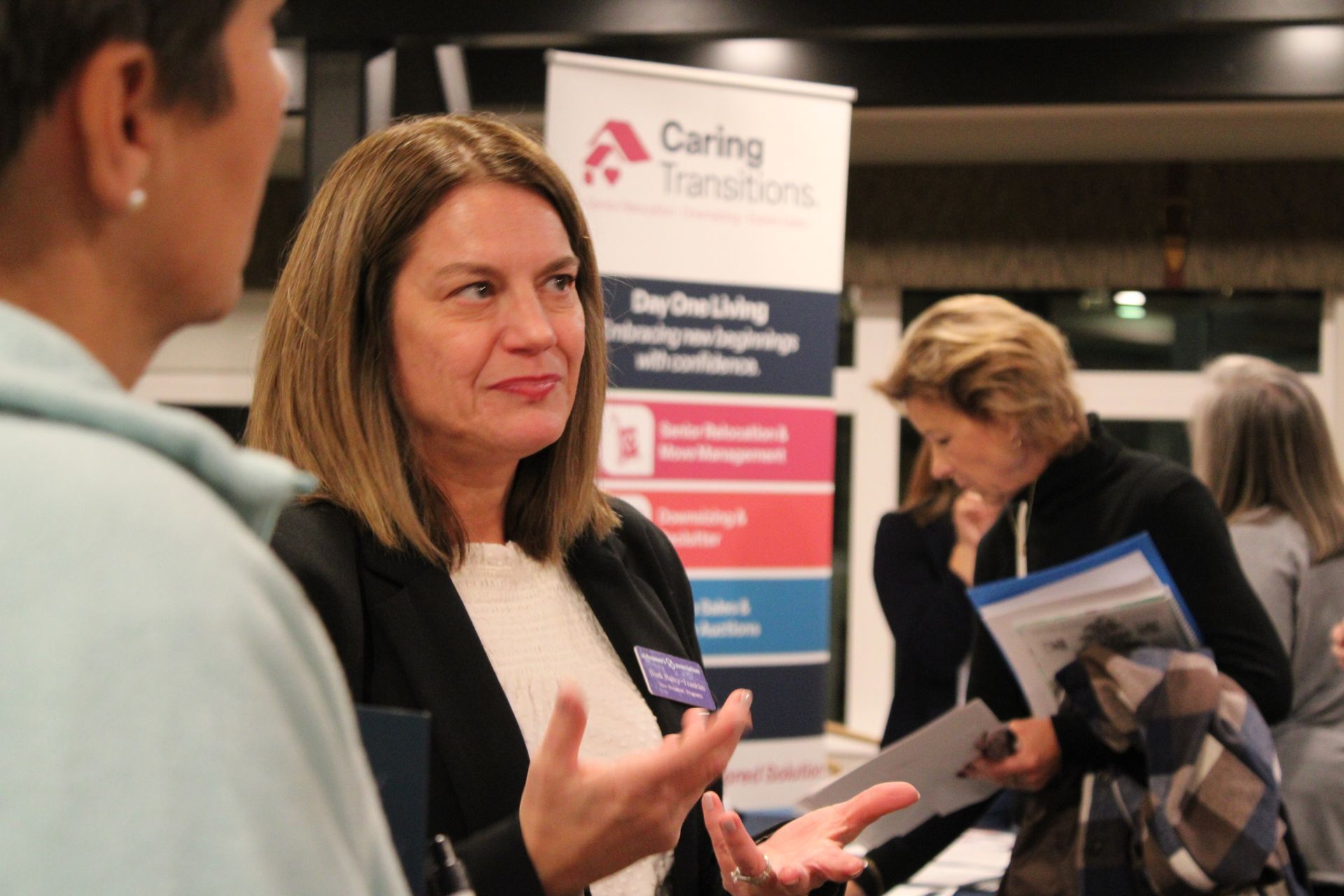 A woman in a black blazer speaks to another person in front of a Caring Transitions banner at an indoor event.