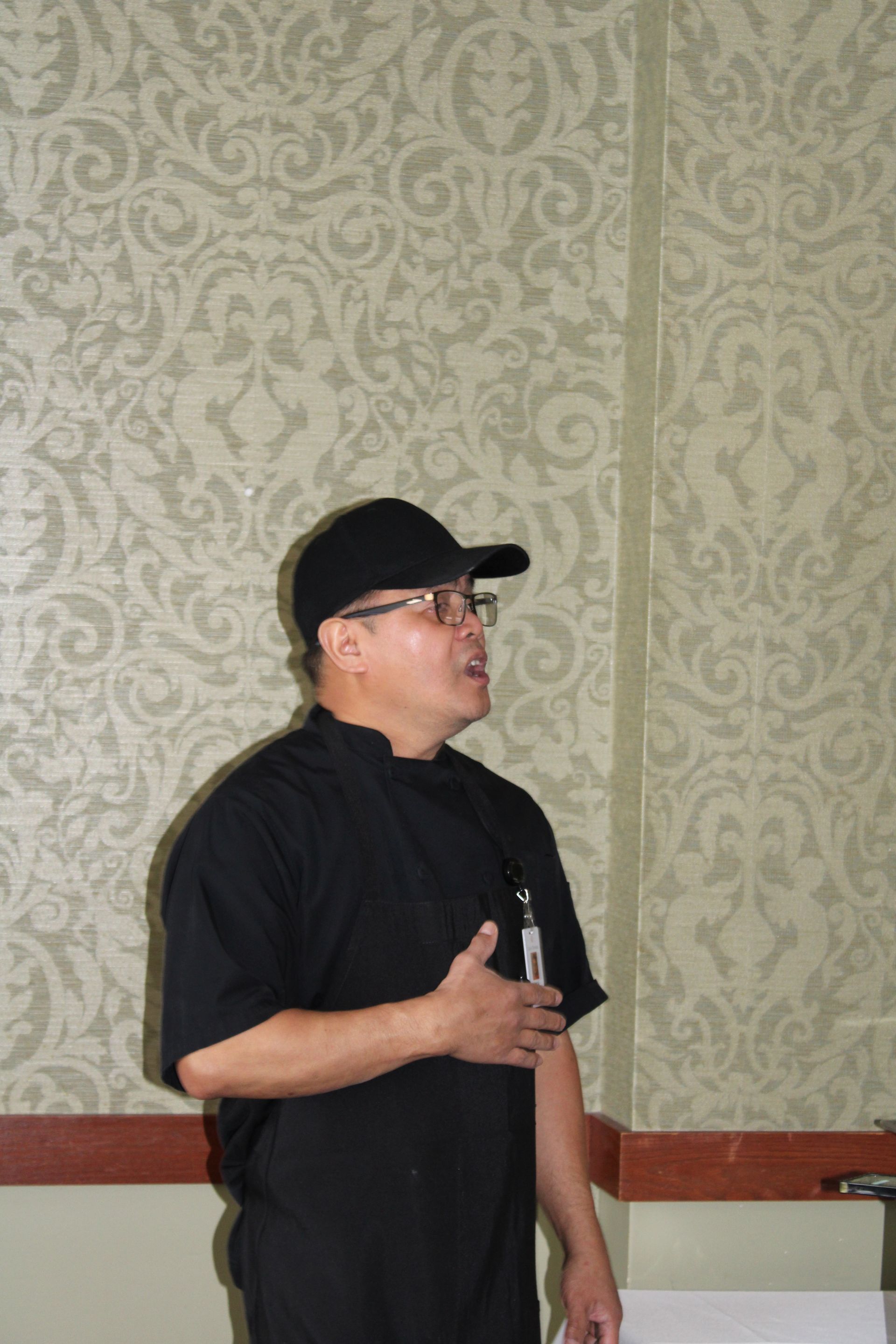 A person in a black cap and shirt standing against a patterned wall, speaking with their hand placed over their chest.