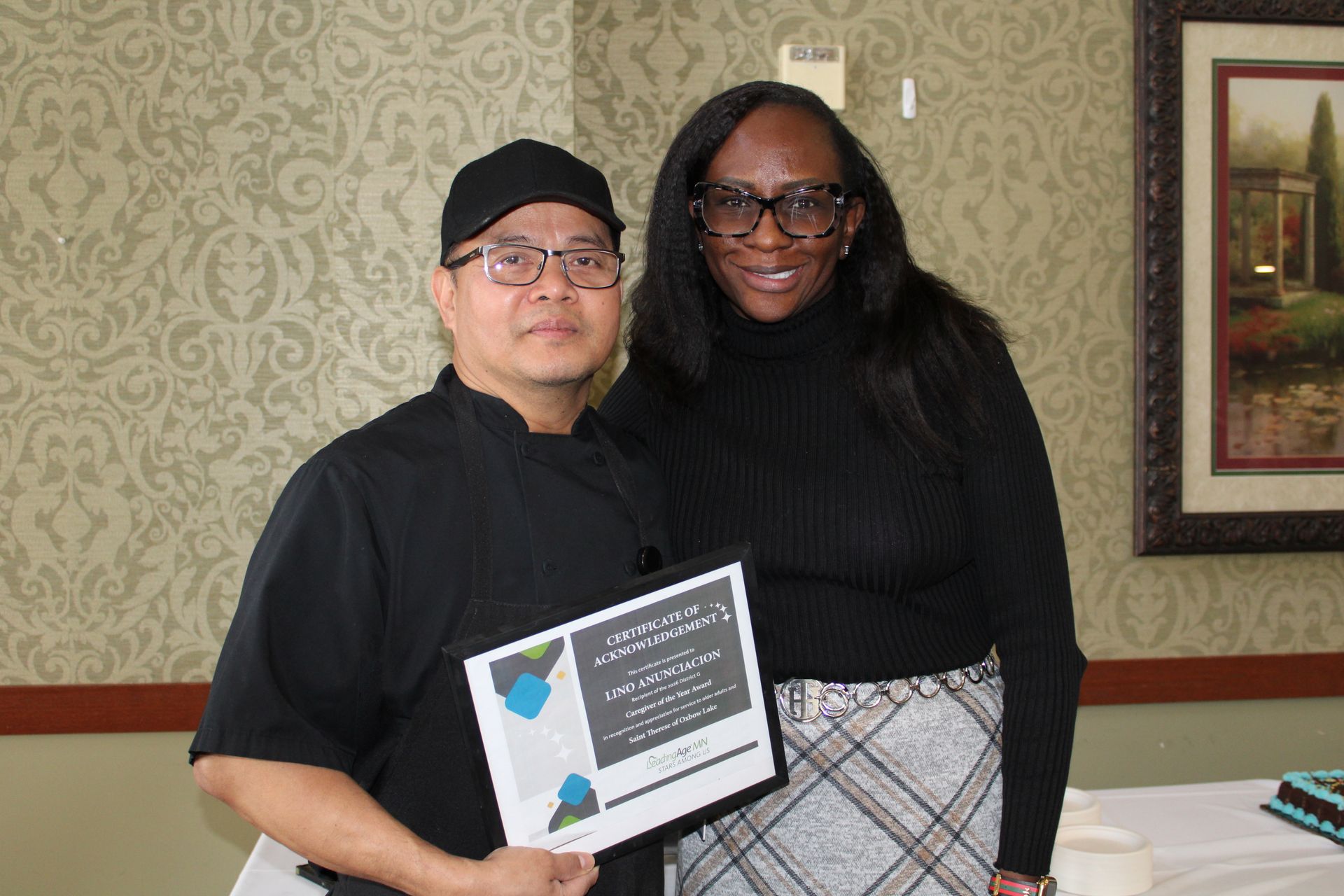 A person in a black chef’s uniform stands with an individual in a black turtleneck, holding an award certificate indoors.