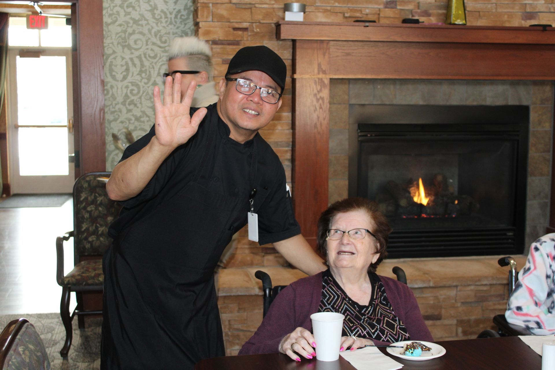 A person in a black chef uniform waves near an older person sitting at a table with a drink, in front of a fireplace.