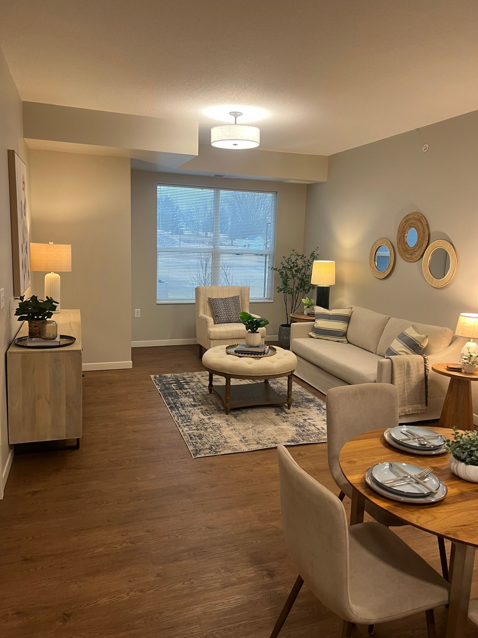 A modern, neutral-toned living and dining area with a beige sofa, patterned rug, arm chair, and a round wooden table.