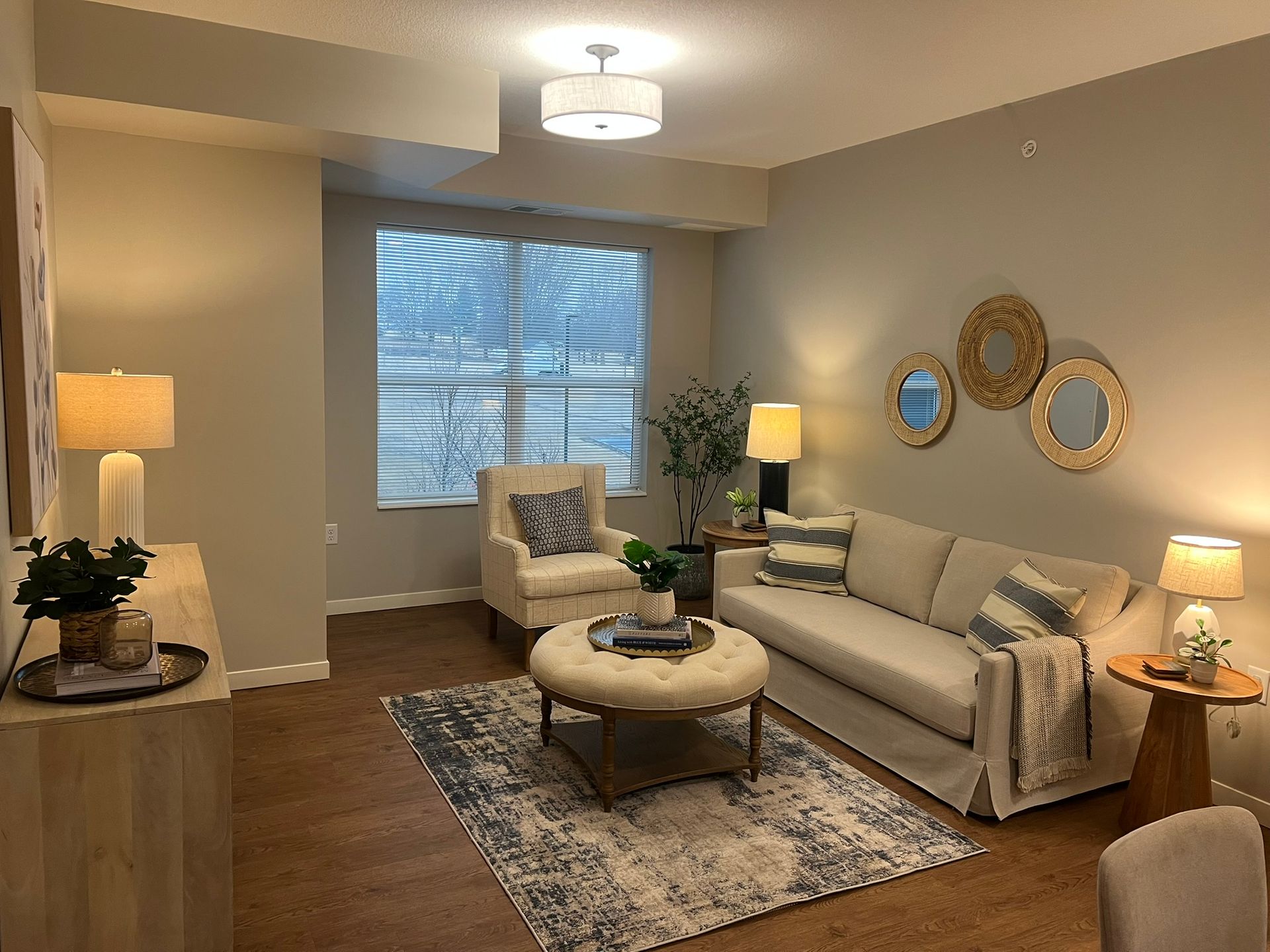 A neutral-toned living room featuring a beige sofa, patterned rug, round ottoman, armchair, and circular wall mirrors.
