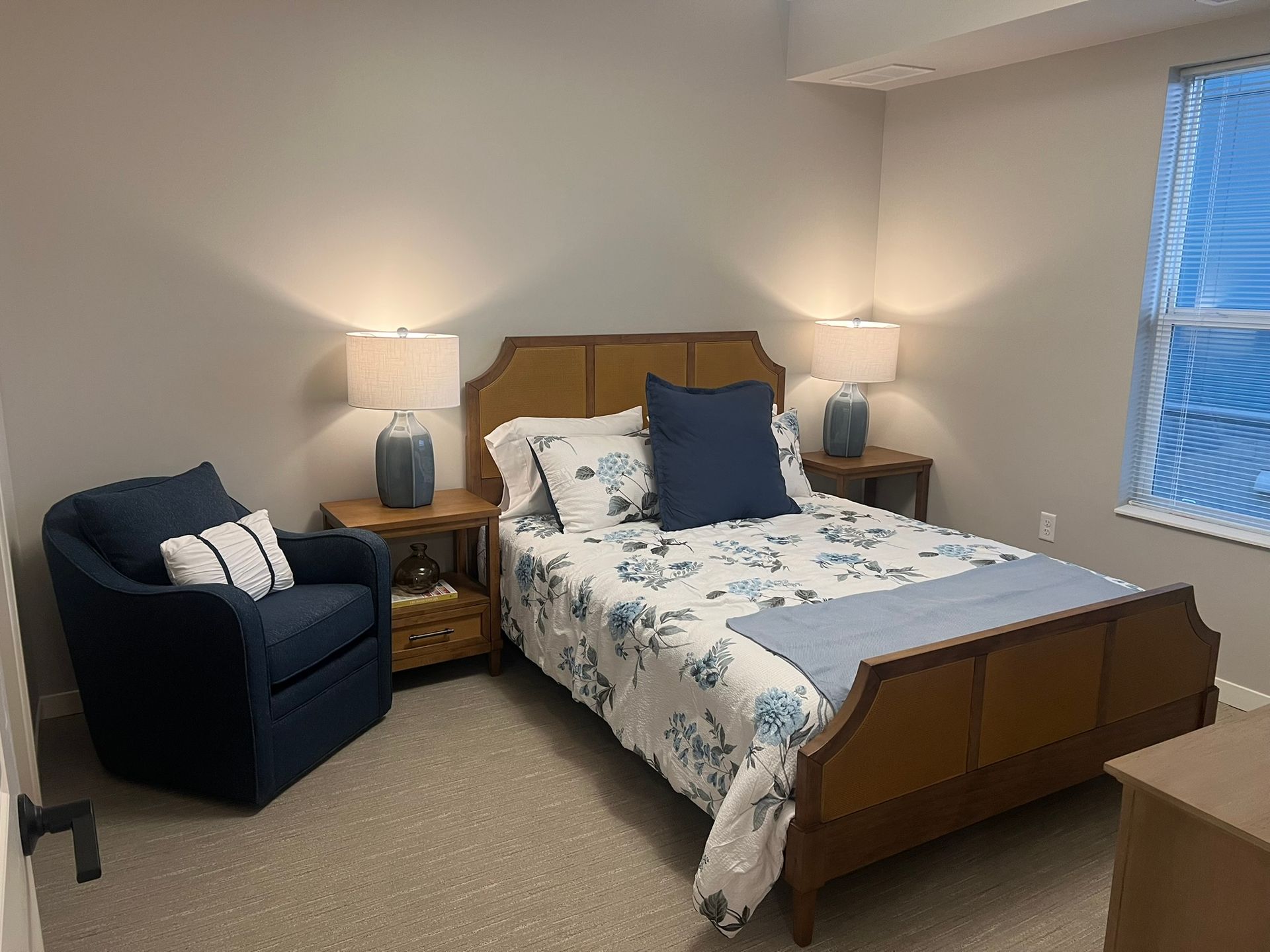 A bedroom with a wooden bed frame, floral bedding, navy accent pillows, a matching navy armchair, and two bedside lamps.