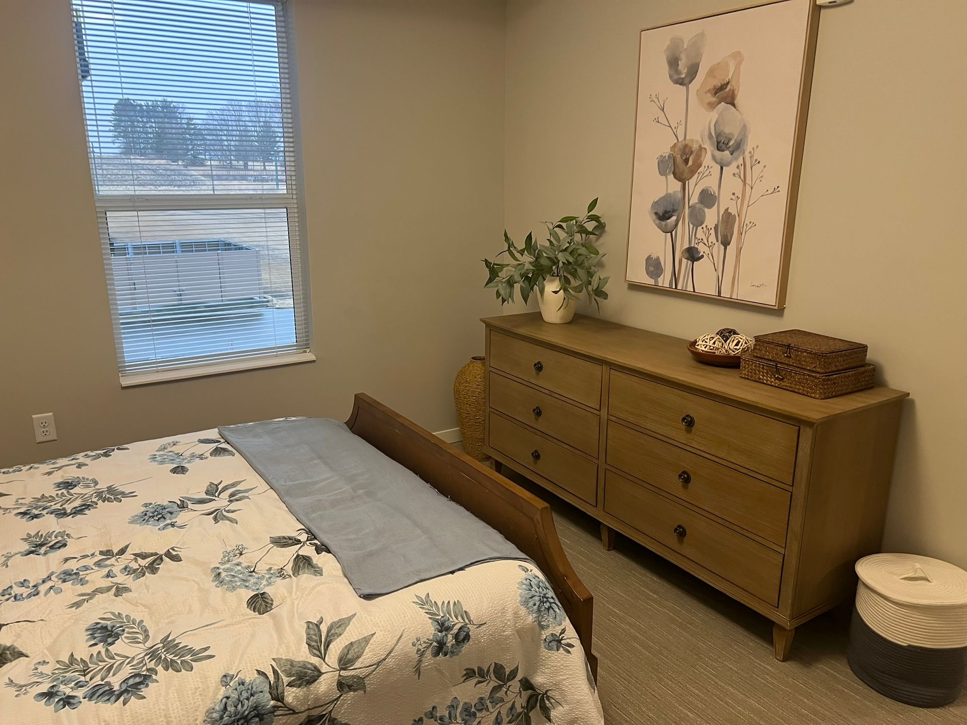 A bedroom featuring a wooden dresser with decor and a bed with a floral blue and white quilt near a window.