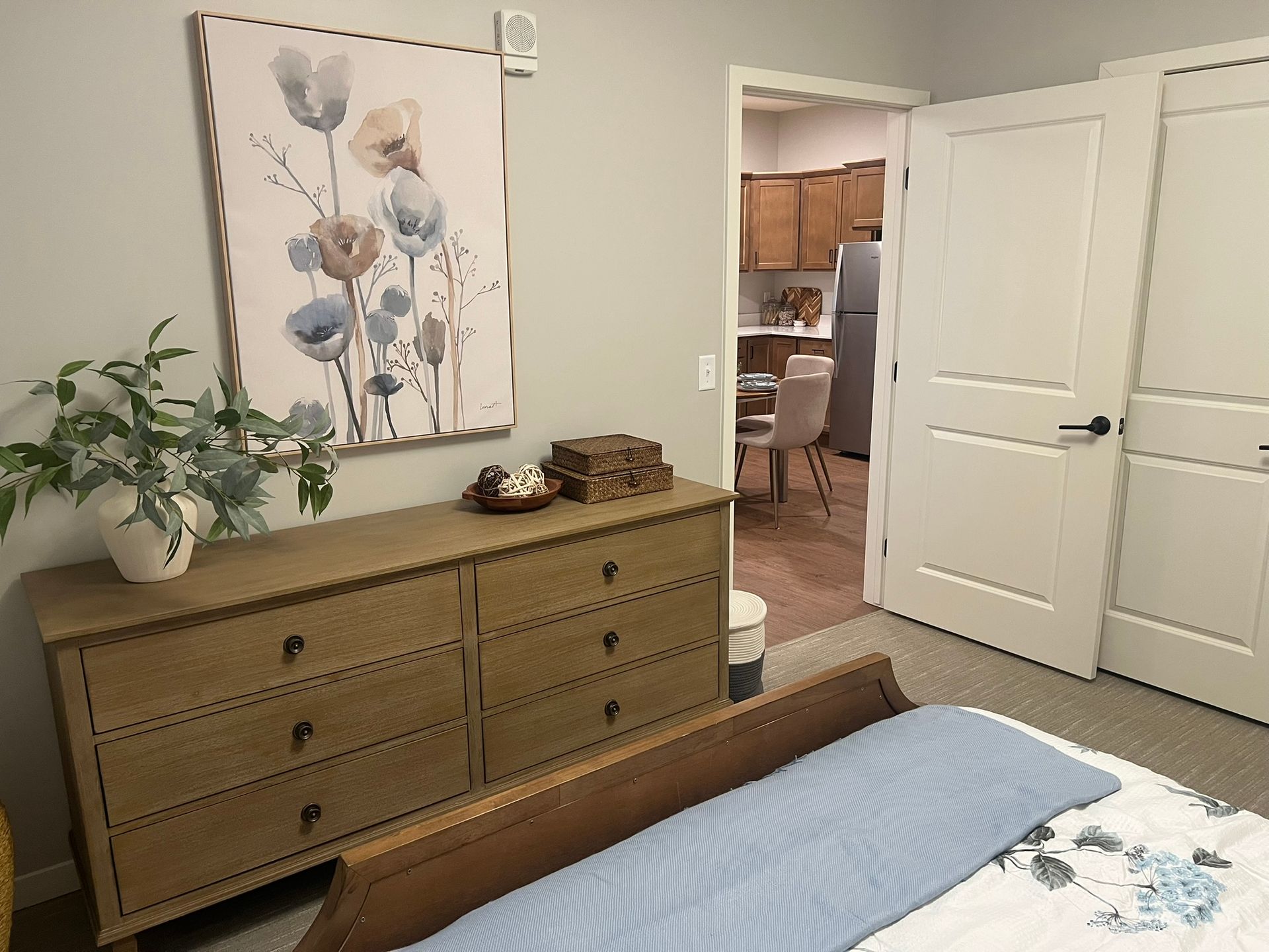 A bedroom featuring a wooden dresser with a plant and artwork, with a door leading to a view of a dining area and kitchen.