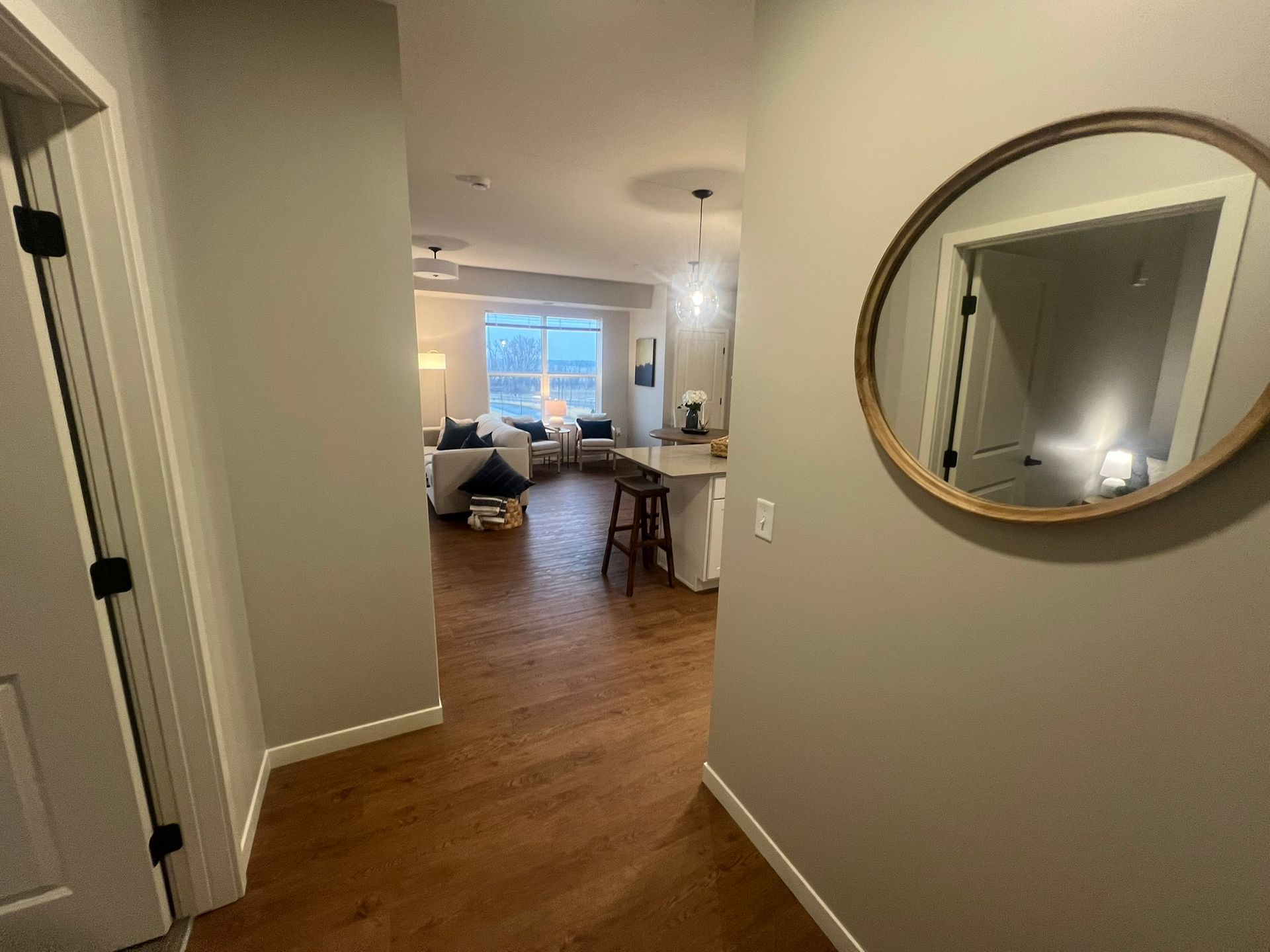 A view through a neutral-toned hallway into an open-plan living space with wooden floors, seating, and a round mirror.