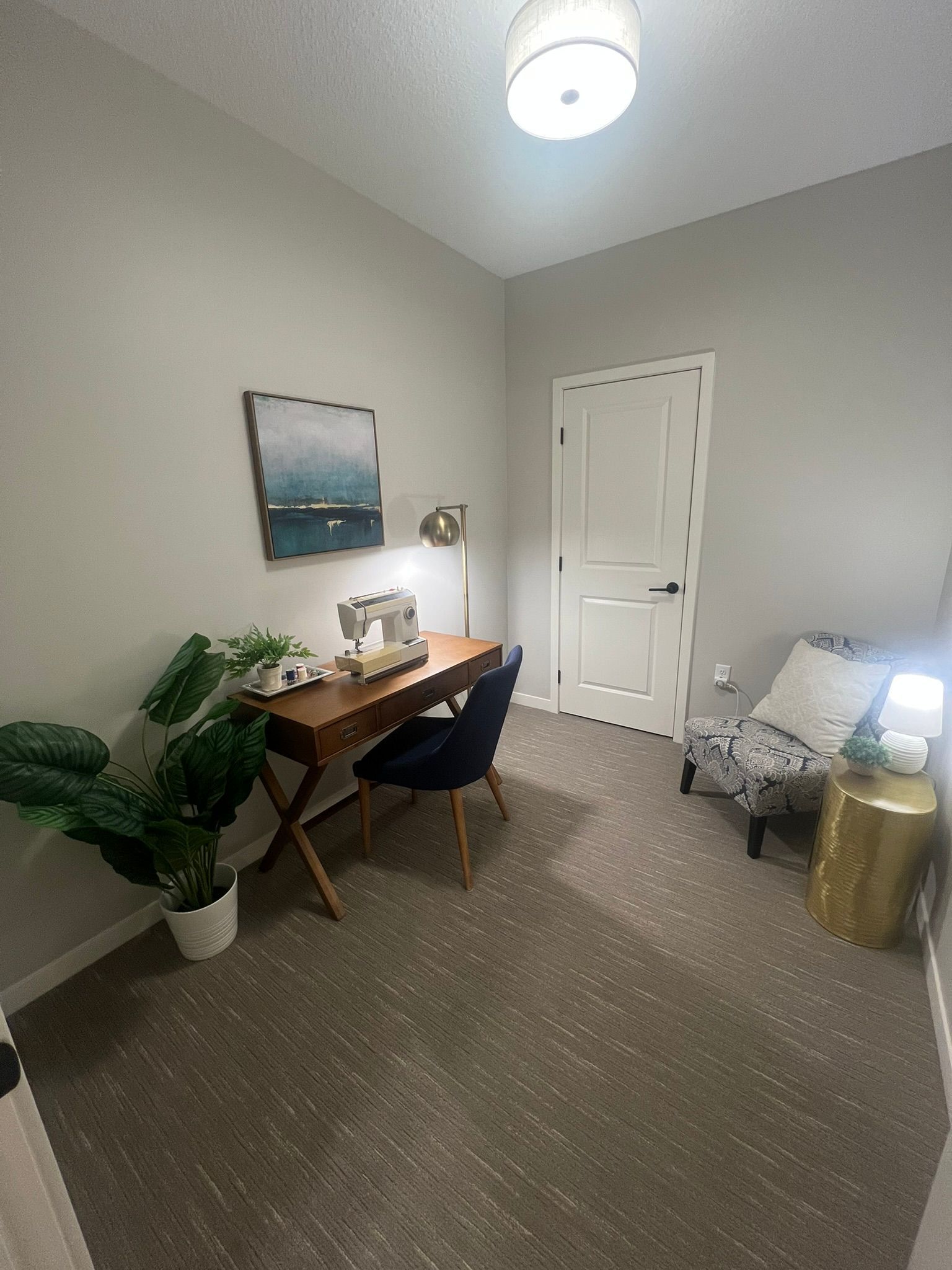 A home office featuring a wooden desk with a sewing machine, a navy chair, a potted plant, and a patterned armchair.