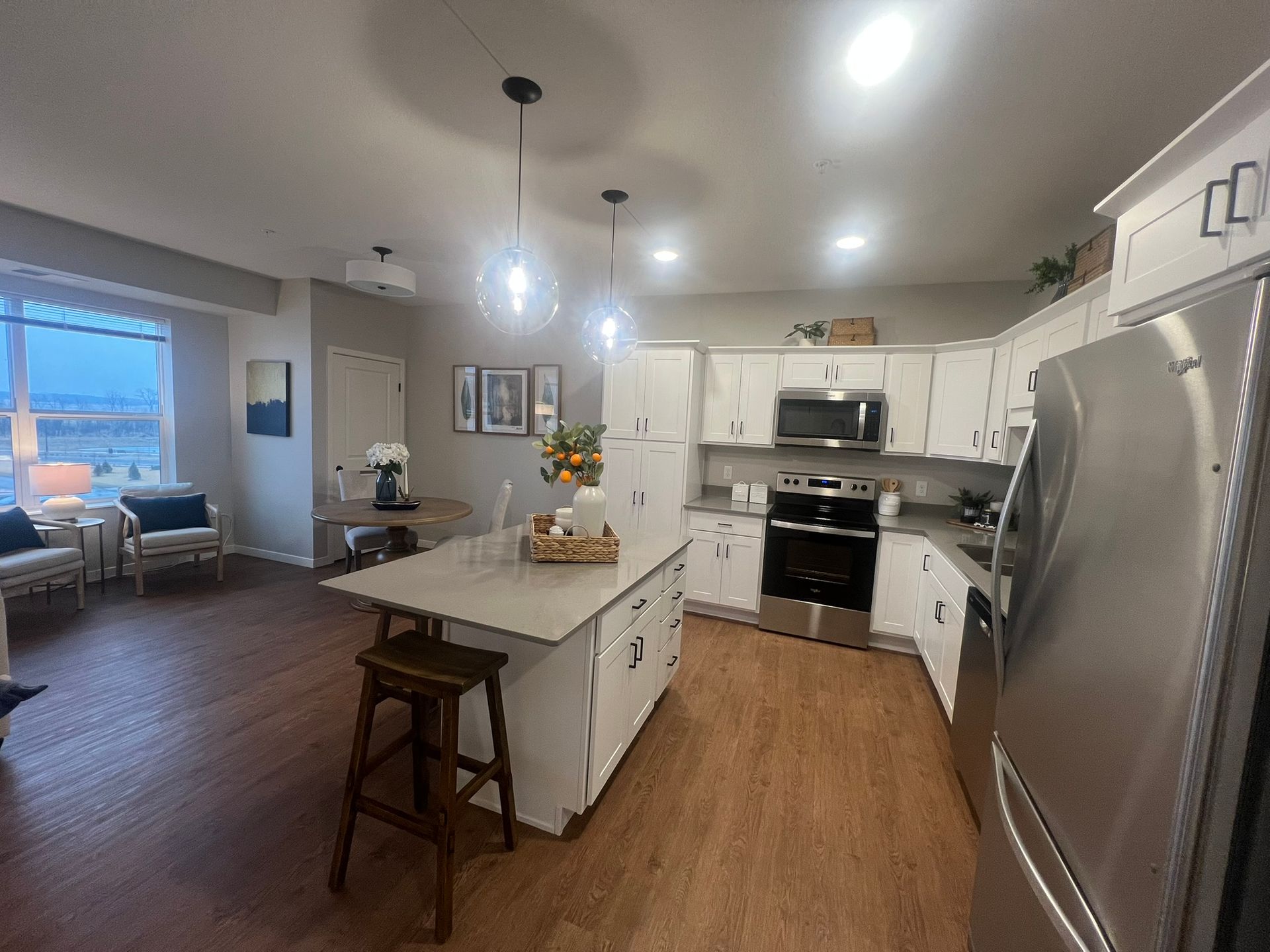 A modern kitchen with white cabinets, a central island with a wooden stool, stainless appliances, and dining area nearby.