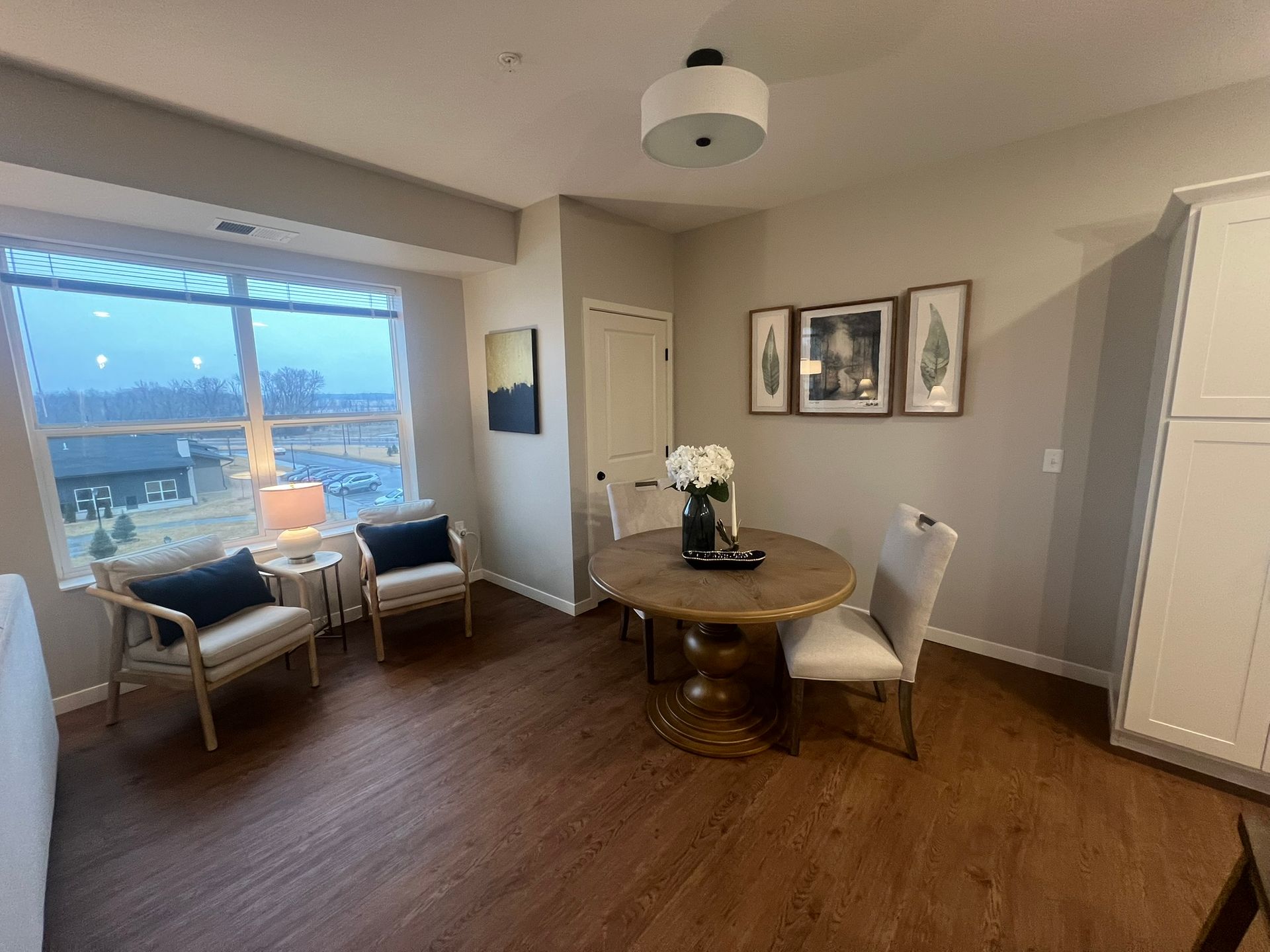 A cozy dining nook featuring a circular wooden table, two chairs, and two armchairs by a large window.