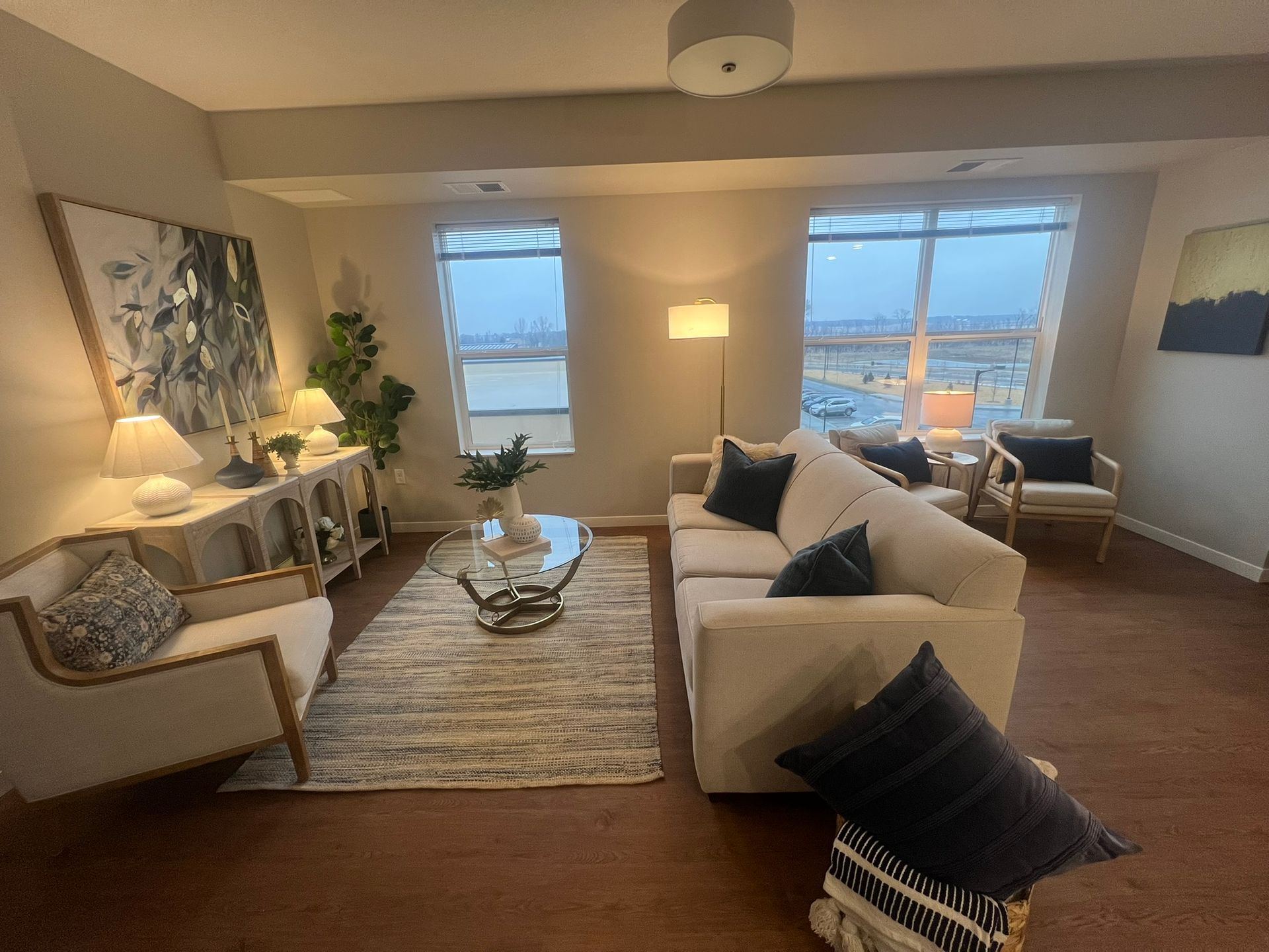 A brightly lit living room with a white sofa, a matching armchair, a glass coffee table, and a rug facing large windows.