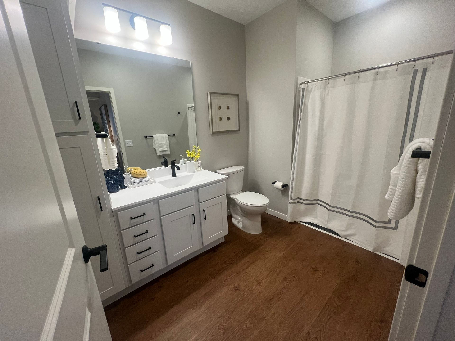 A bright bathroom featuring white vanity cabinets, a large mirror, a toilet, and a shower with a white curtain.