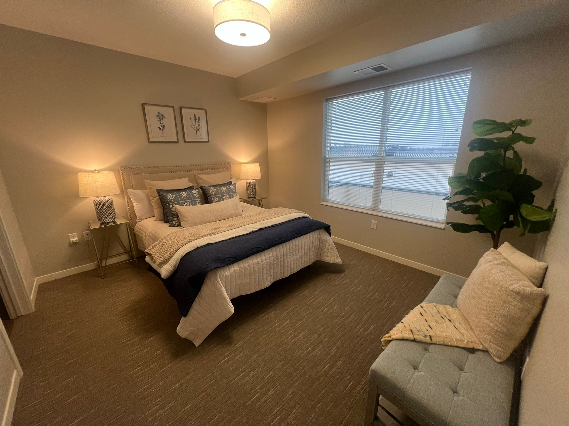 A cozy, neutral-toned bedroom with a queen bed, two lamps, wall art, a large window, and a bench at the foot of the bed.