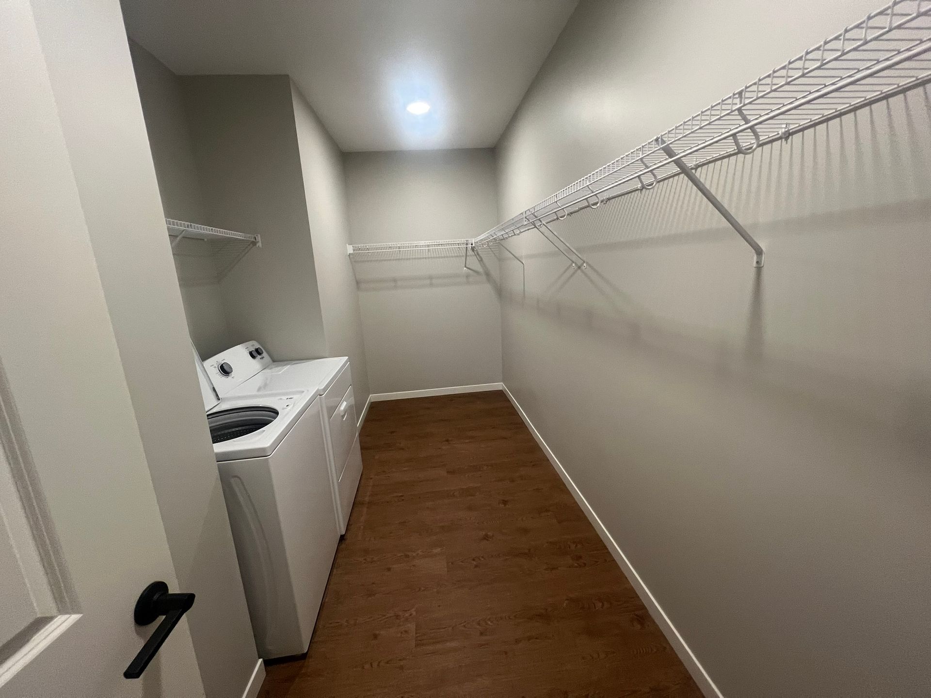 A laundry closet with white appliances, wood-look flooring, and wall-mounted wire shelving.