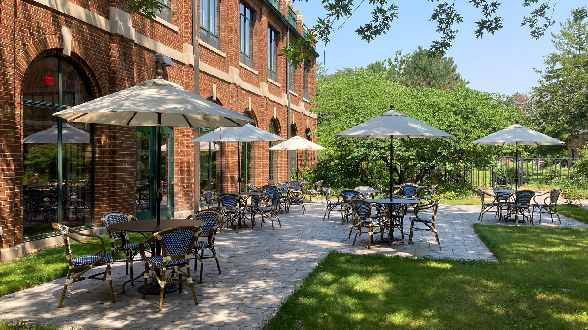 Outdoor patio with tables and umbrellas set against a brick building facade on a sunny day.