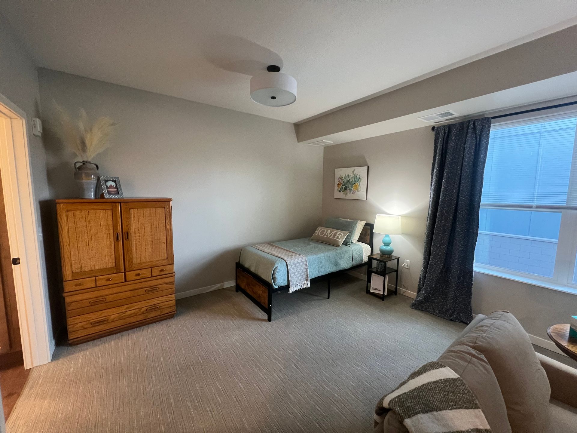 A simple bedroom with a wooden armoire, a single bed with light blue bedding, a nightstand, and gray curtains.
