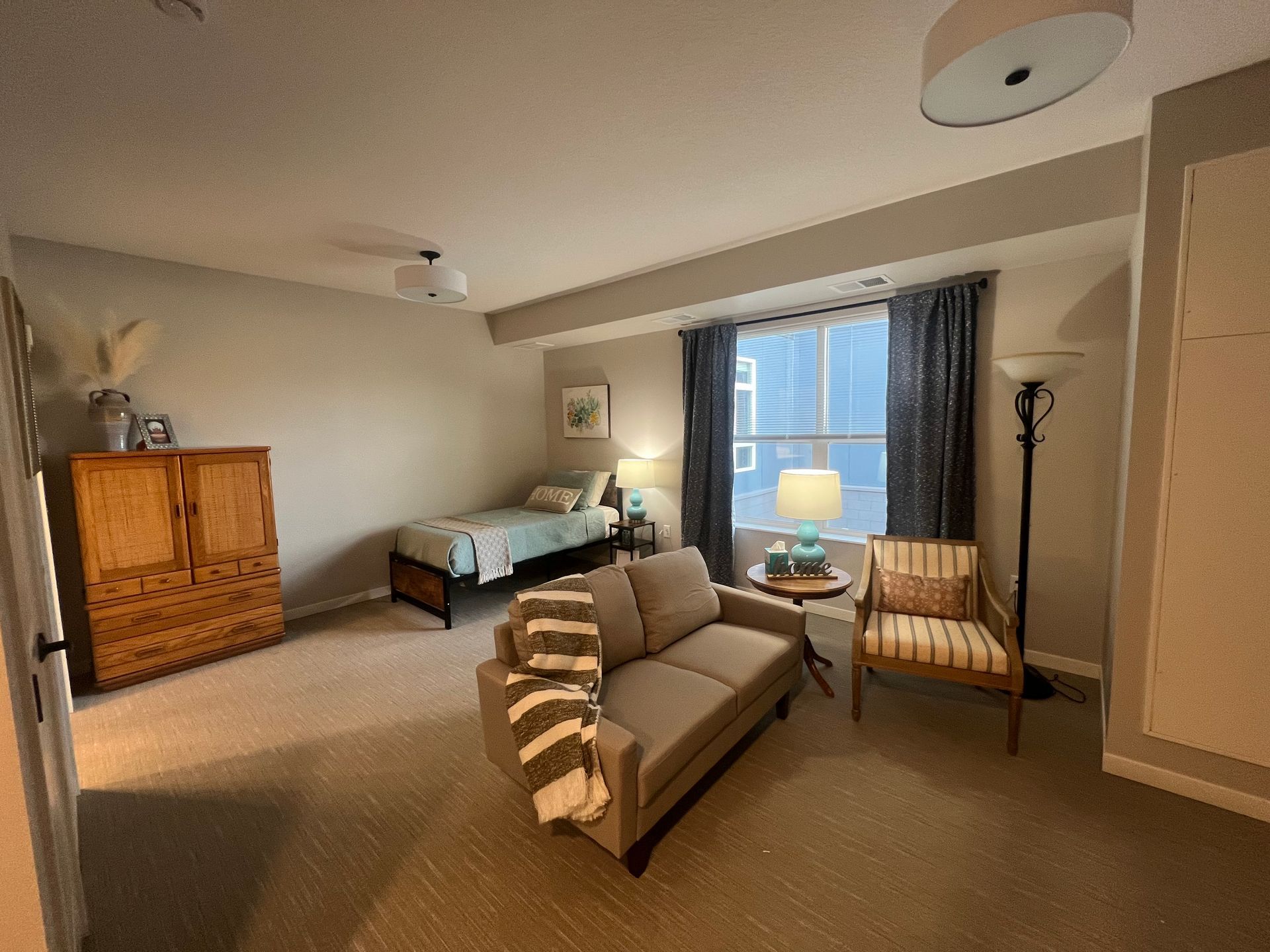 A bedroom with a wooden dresser, a twin bed, a sofa with a striped throw, a patterned armchair, and a floor lamp.