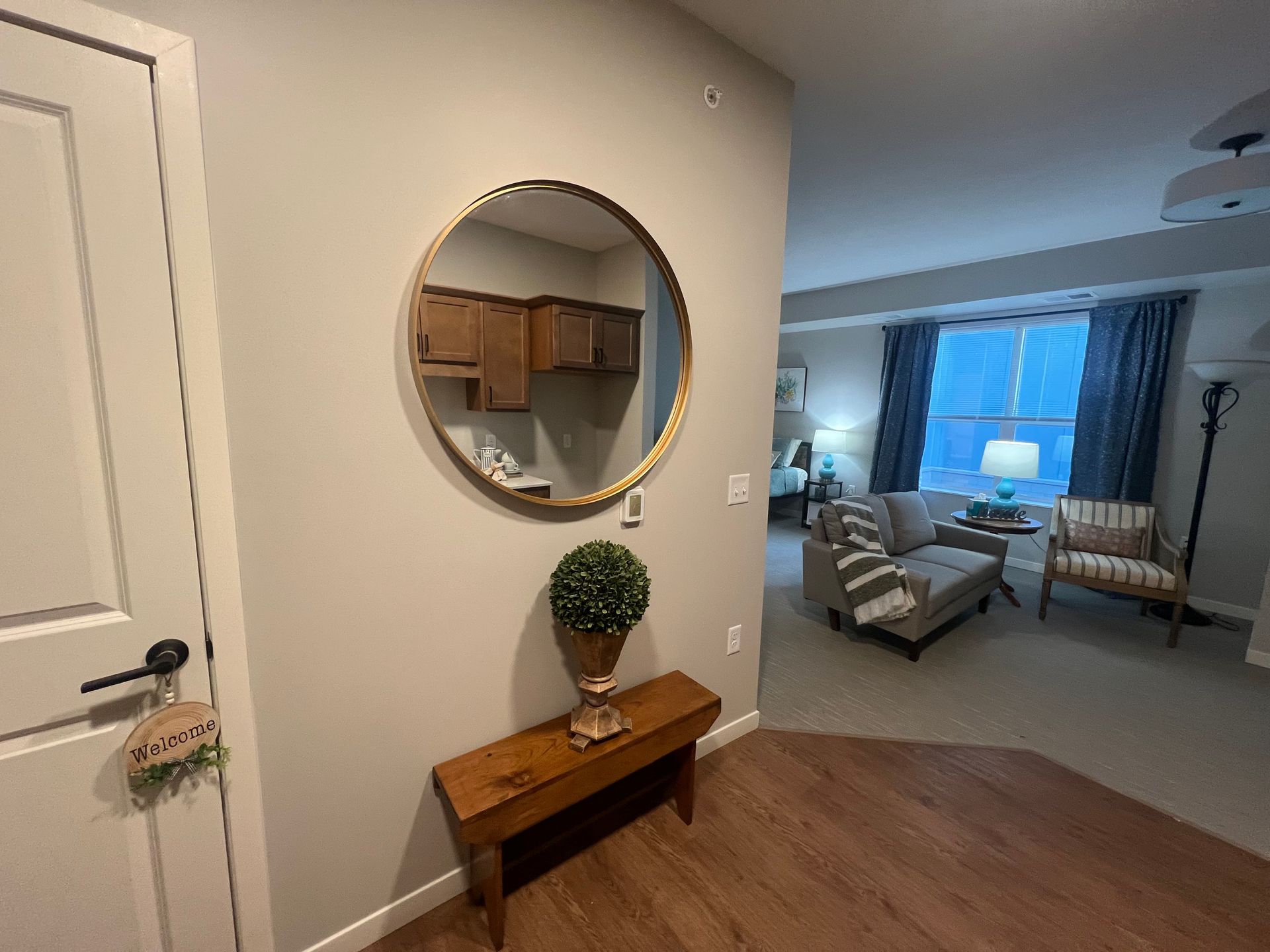 A entryway with a wood bench and potted plant under a round mirror, looking into a living room with sofa and armchair.