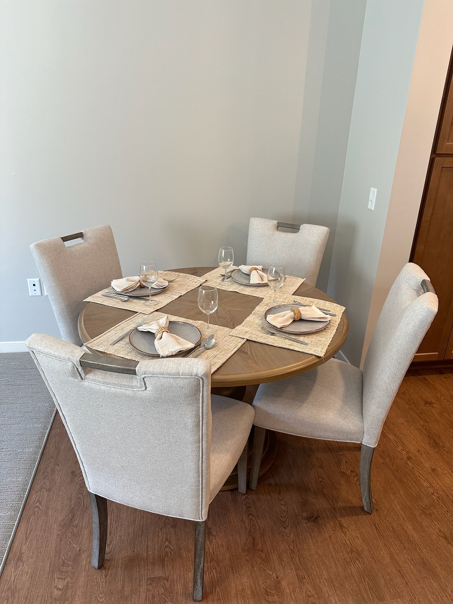 A round wooden table with four beige upholstered chairs in a dining area, set with place mats, plates, and wine glasses.