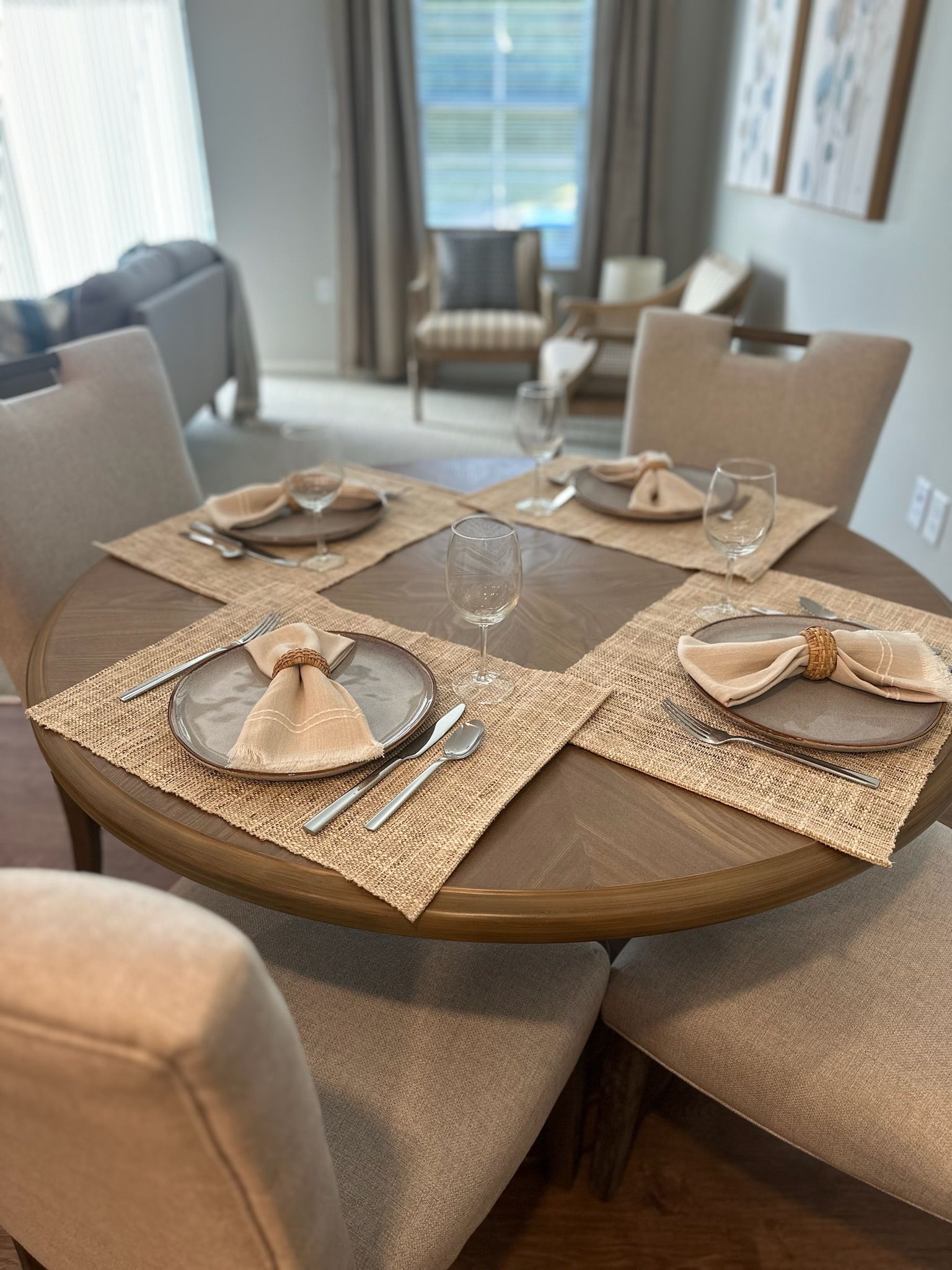 A round wooden dining table set for four with woven placemats, gray plates, cream-colored napkins, and wine glasses.