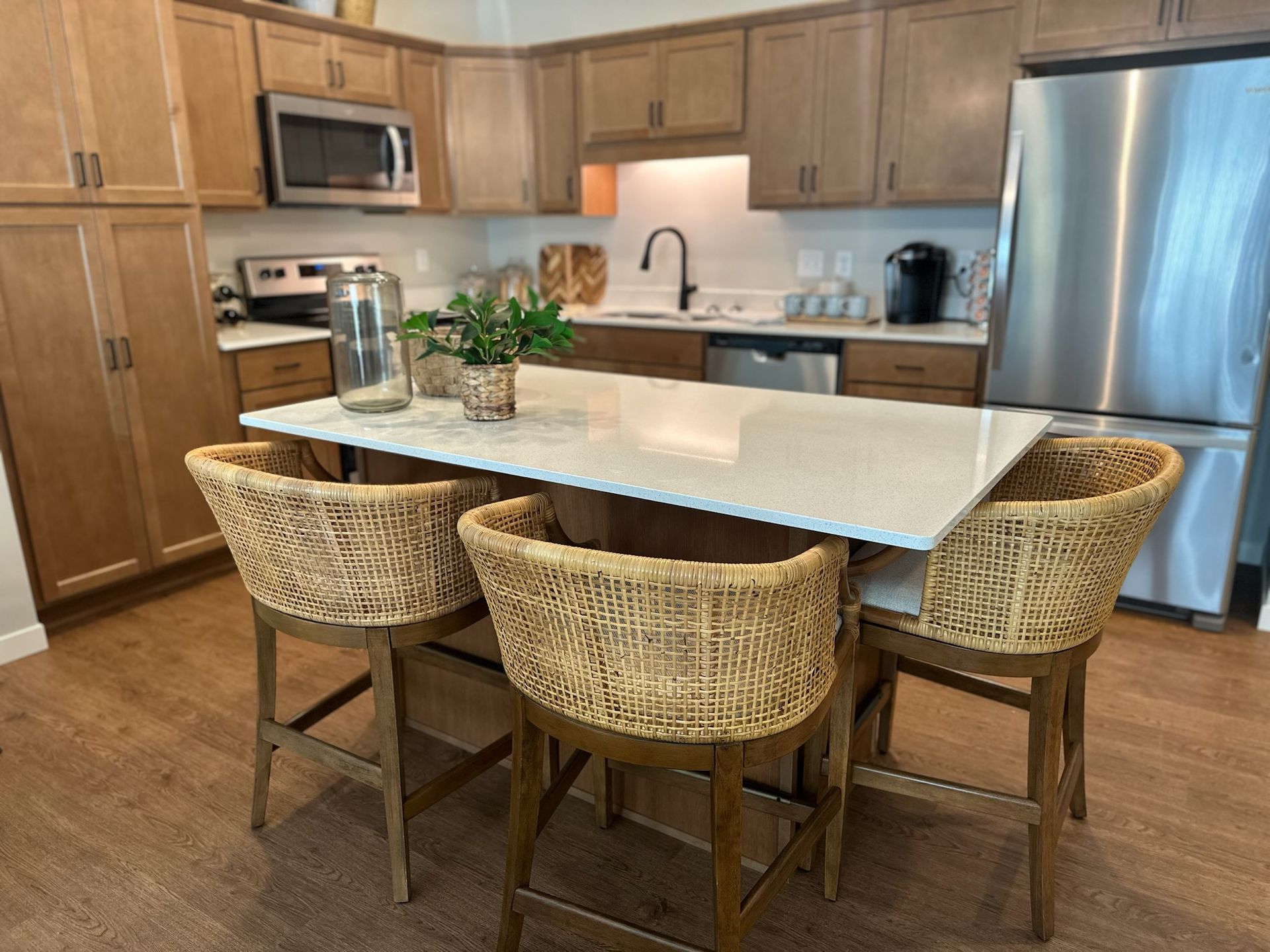 A modern kitchen with light wood cabinets, stainless steel appliances, and a center island with three woven bar stools.