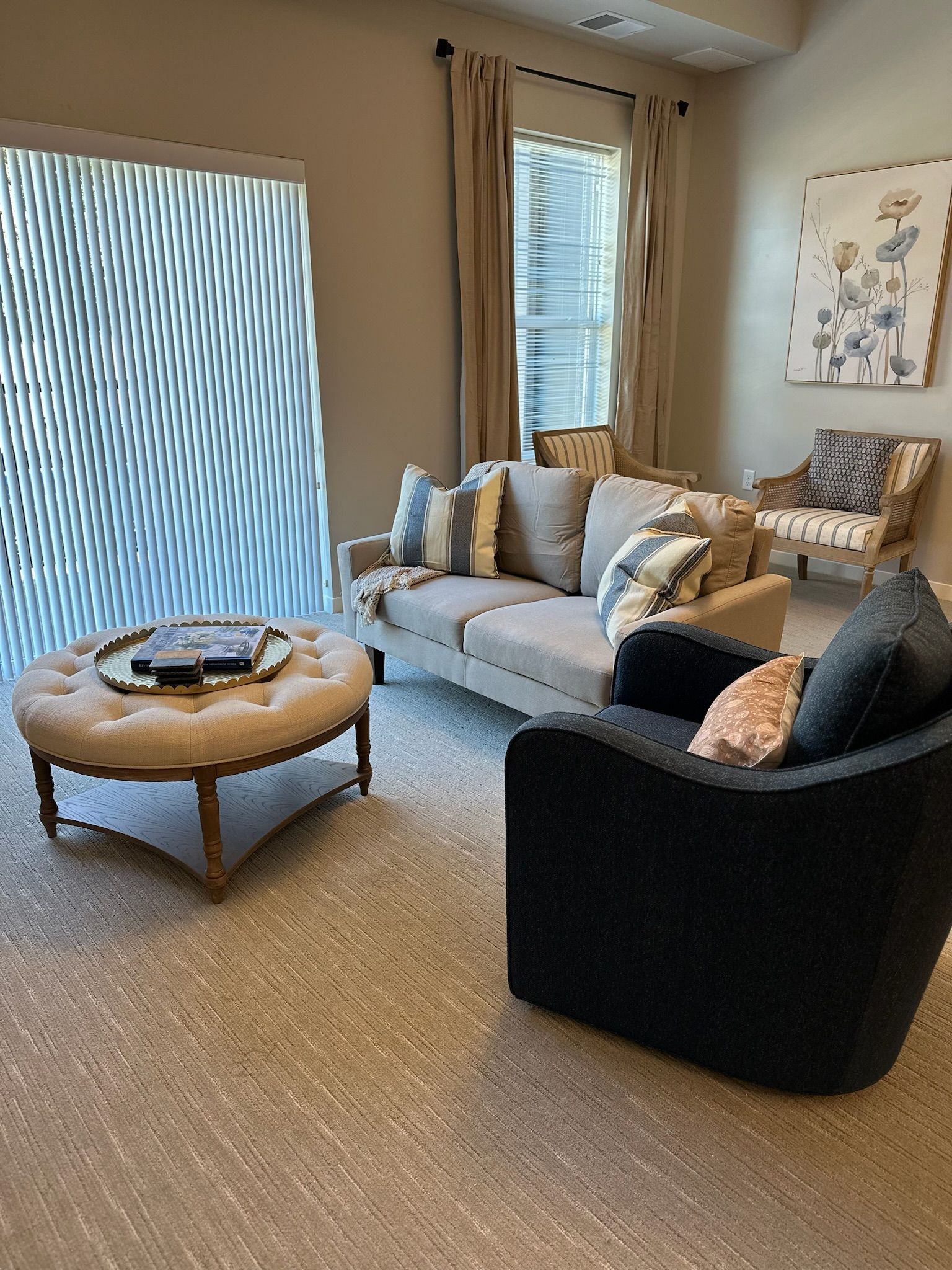 A cozy living room with a neutral-toned sofa, a round tufted ottoman, a dark armchair, and an accent chair near a window.